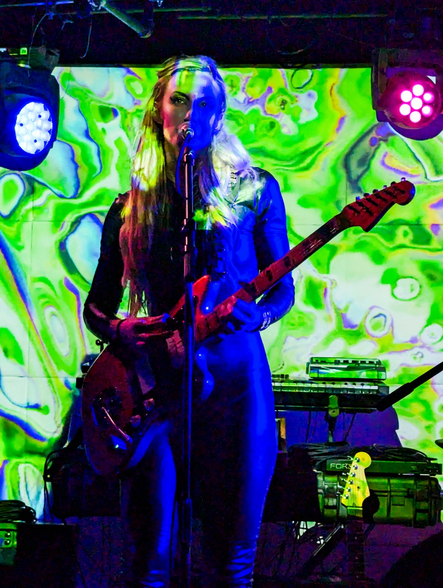 Female musician playing electric guitar on stage with colorful abstract background, stage lights, and equipment.
