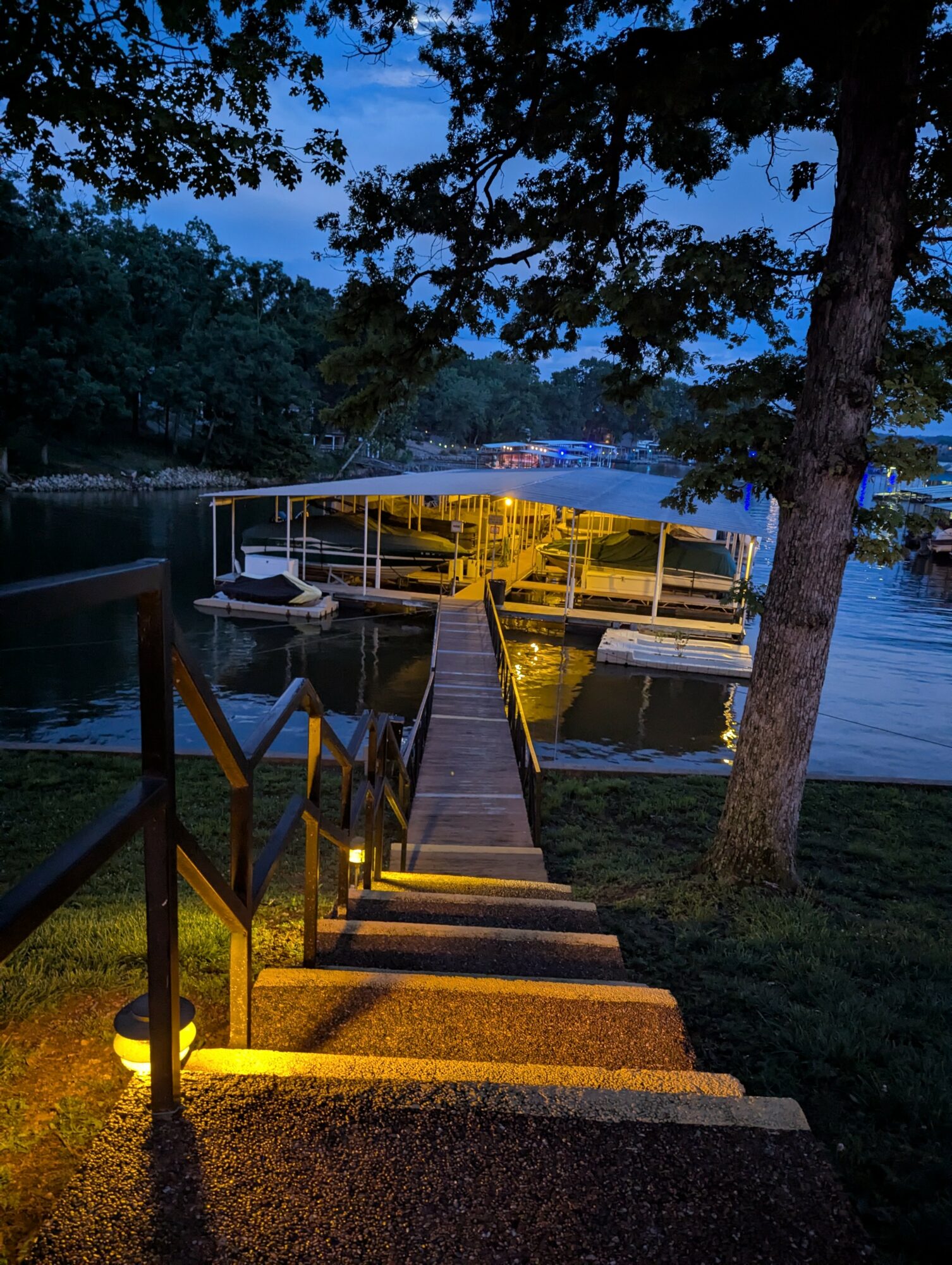 Staircase leading to a dock with boats on a lake at dusk, illuminated by yellow lights, trees on both sides.