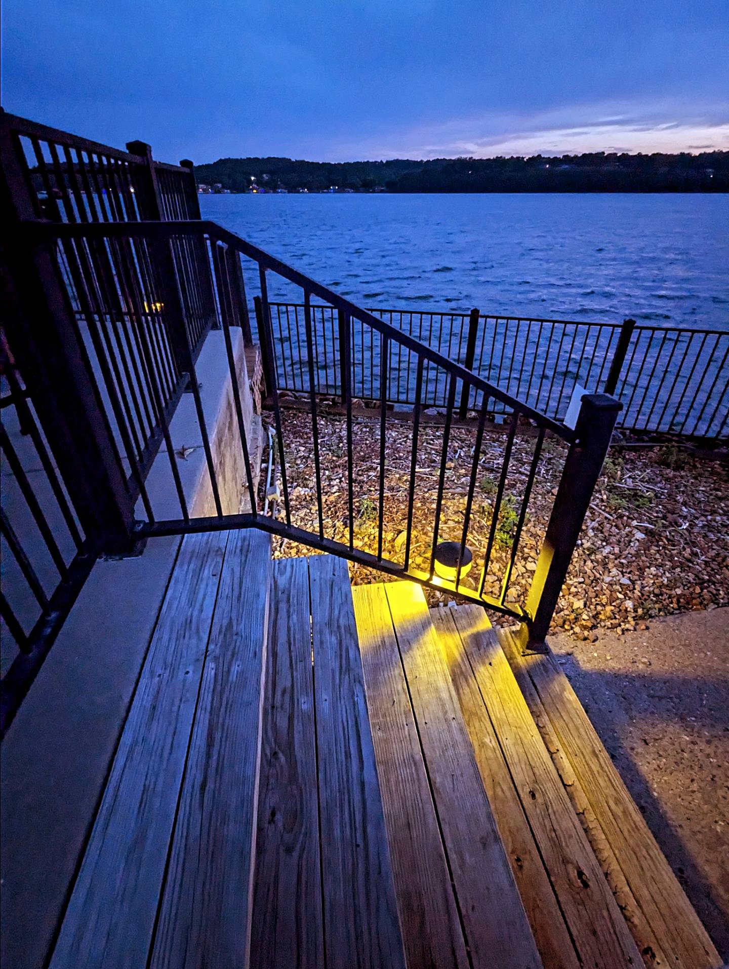 Staircase leading to a waterfront with a metal railing, illuminated by yellow lights, under a blue evening sky.