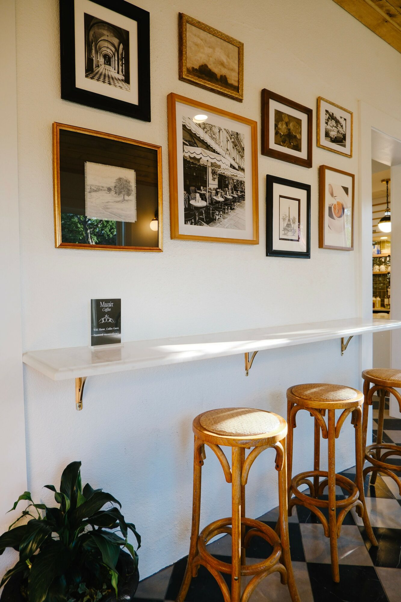 Wall with framed pictures above a white counter, two wooden stools, and a potted plant in a cafe or gallery setting.