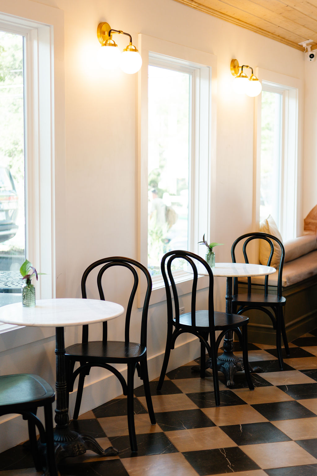 Interior of a cafe with white walls, large windows, black chairs, and checkered floor, illuminated by wall-mounted lights.