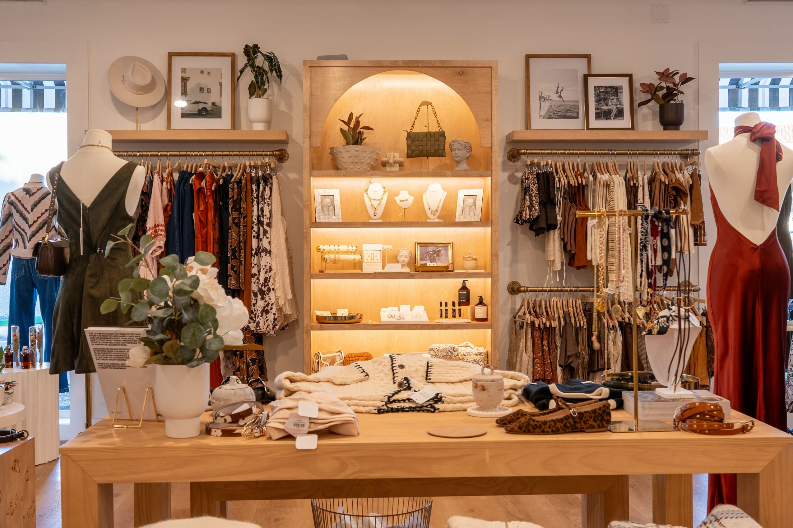 Clothing store display with mannequins, shelves of accessories, and a wooden table with folded clothes and flowers.