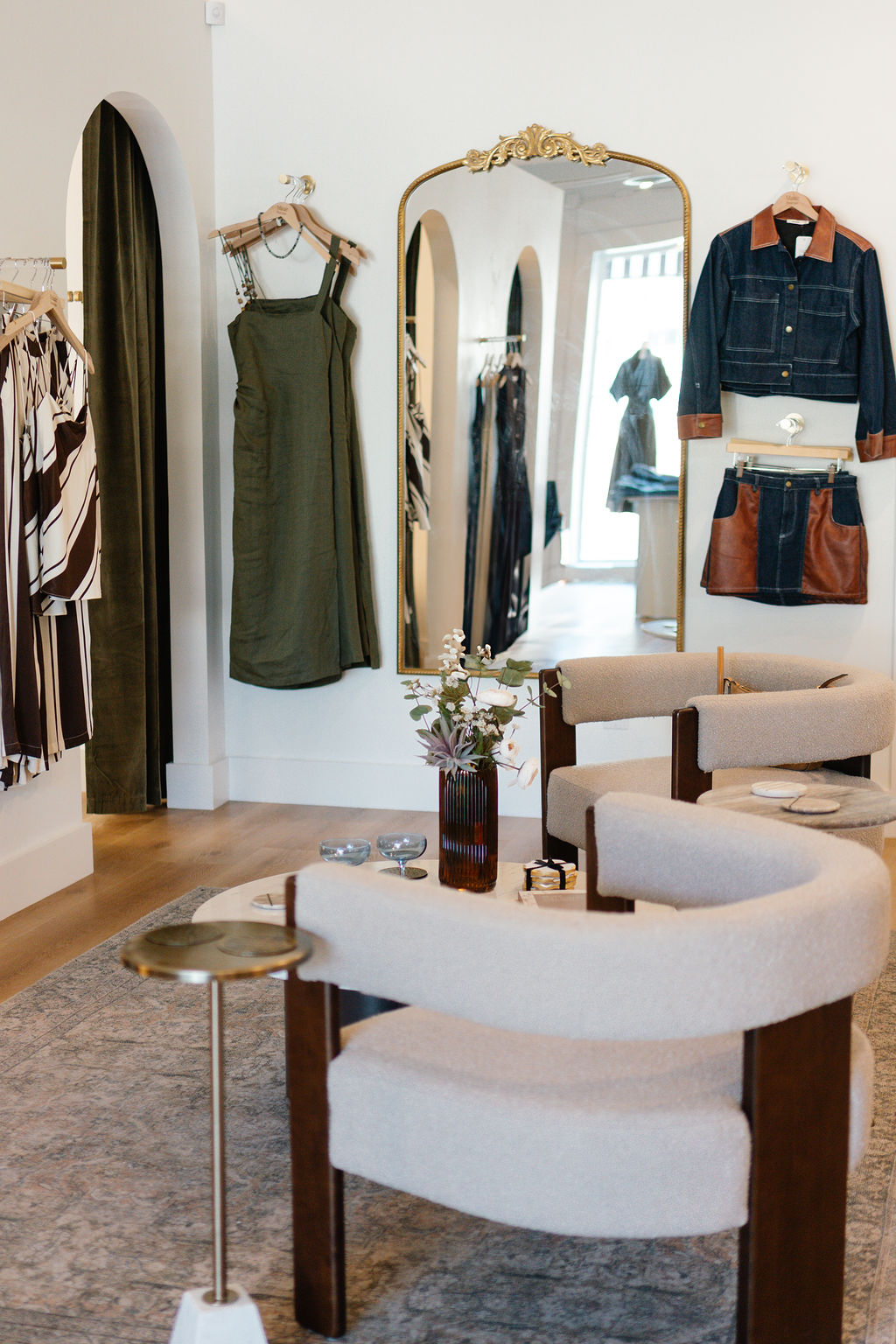 Clothing store interior with dresses, a mirror, and a seating area with a vase of flowers.