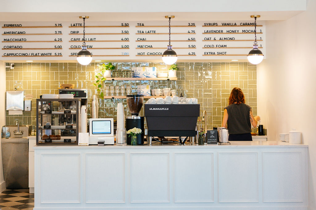 Coffee shop counter with a barista, menu above, and equipment behind the counter, with a person facing away. Bright and modern.