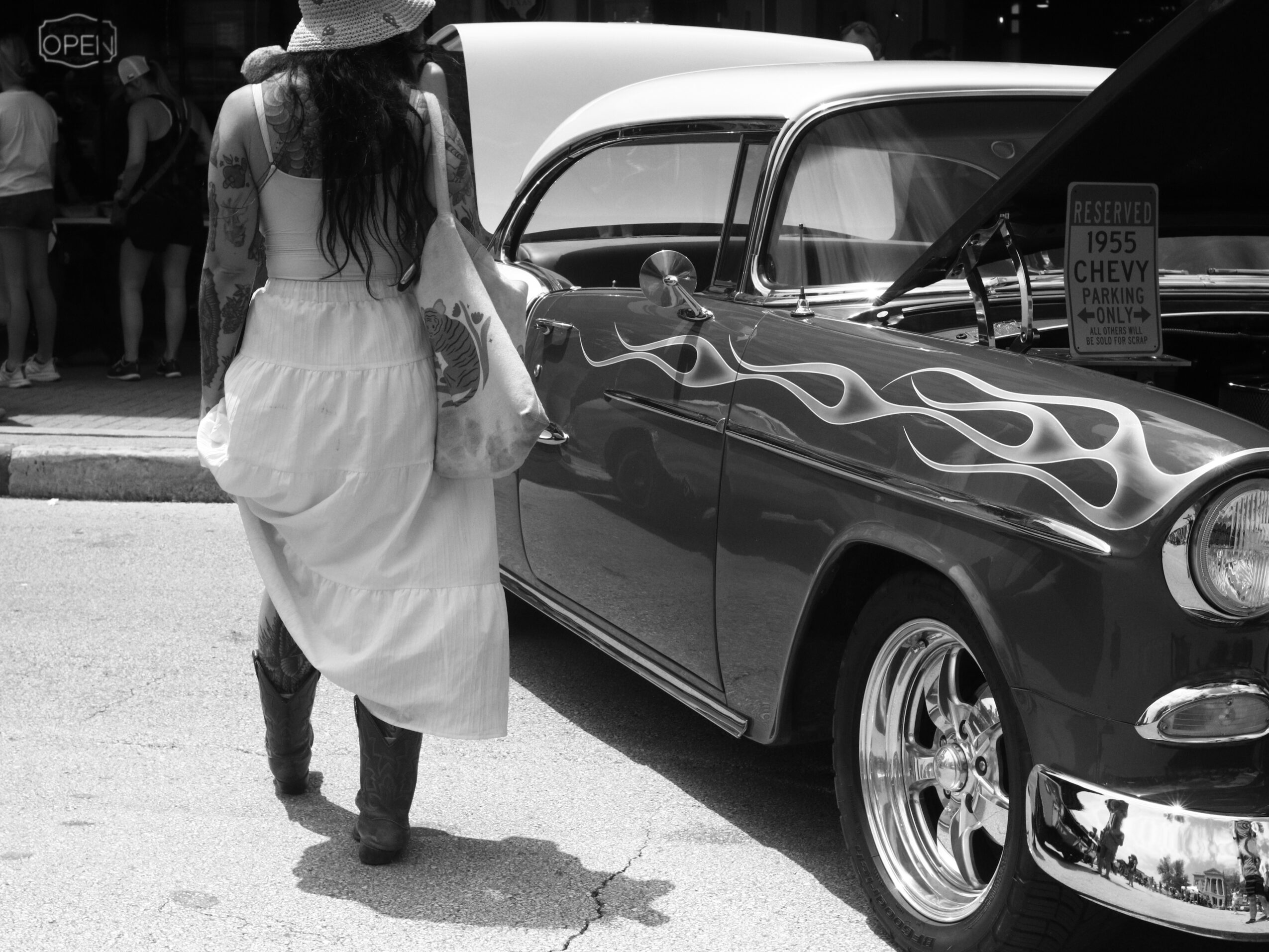 Woman in a white dress and hat walking past a vintage car with flame decals, parked outdoors.