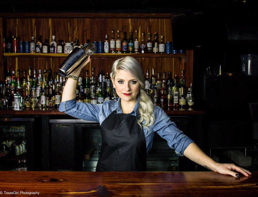 Woman bartender with blonde hair, wearing a black apron and blue shirt, shaking a cocktail shaker behind bar.