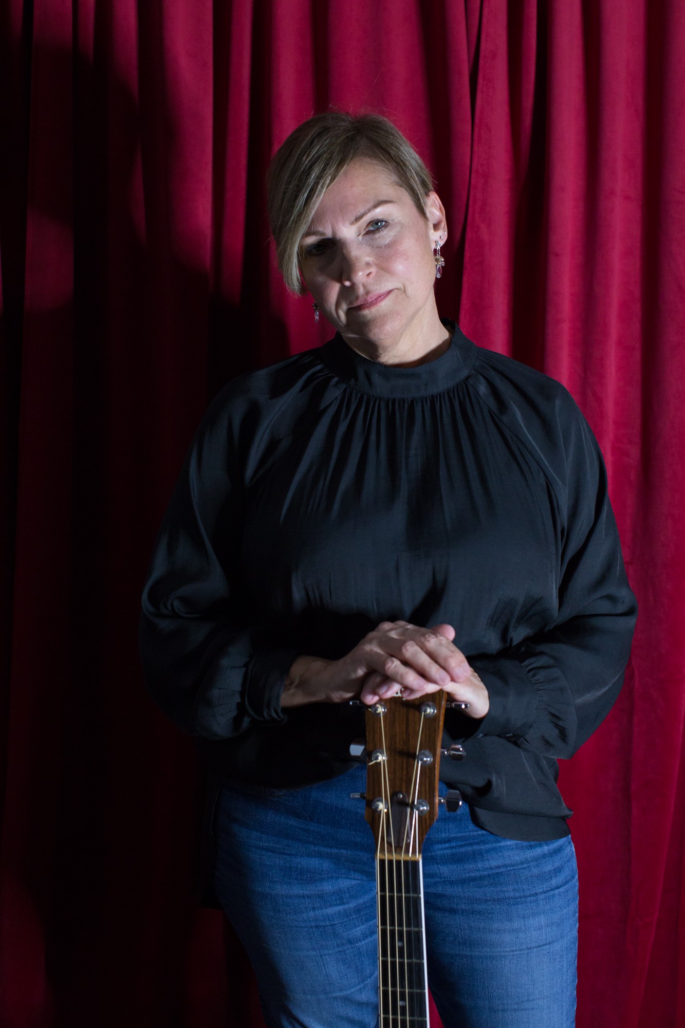 Woman with short hair in a black top standing in front of red curtains, holding a guitar.