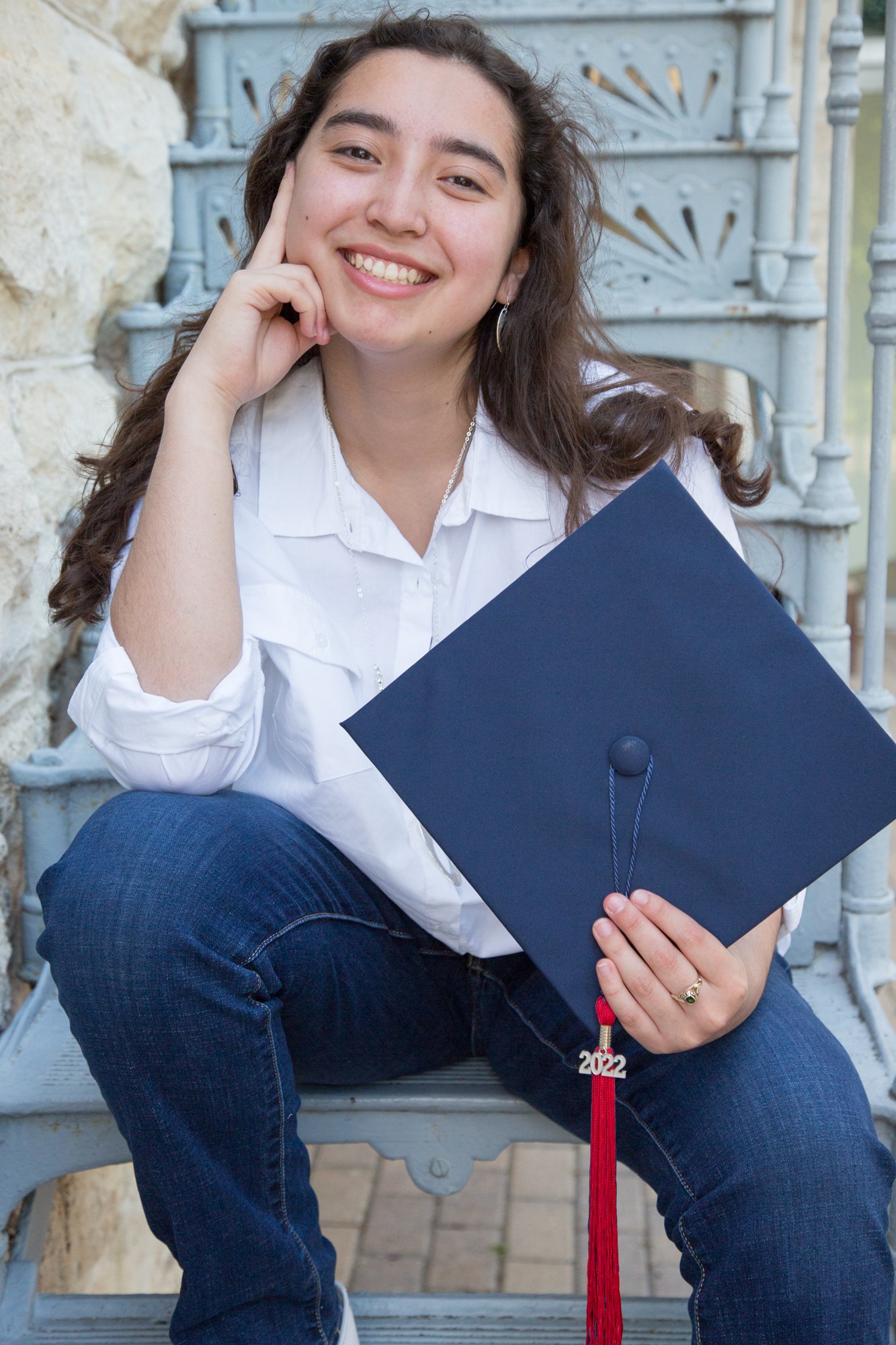 Young woman smiling, holding a graduation cap, sitting outdoors in front of a decorative gate.