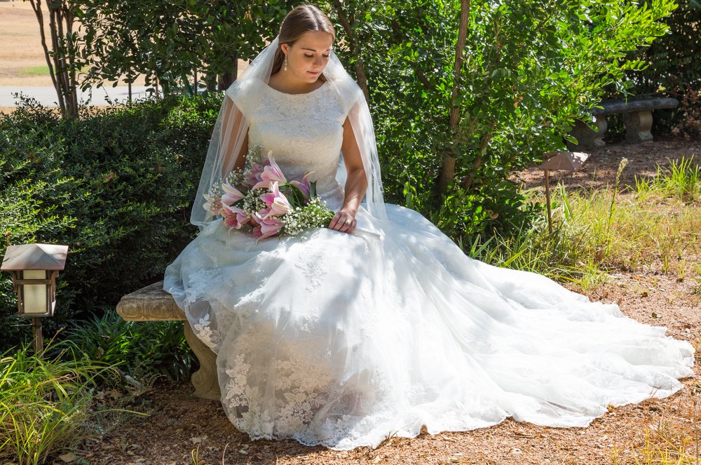 Bride in a white wedding dress sitting on a bench outdoors, holding a bouquet, surrounded by greenery and sunlight.
