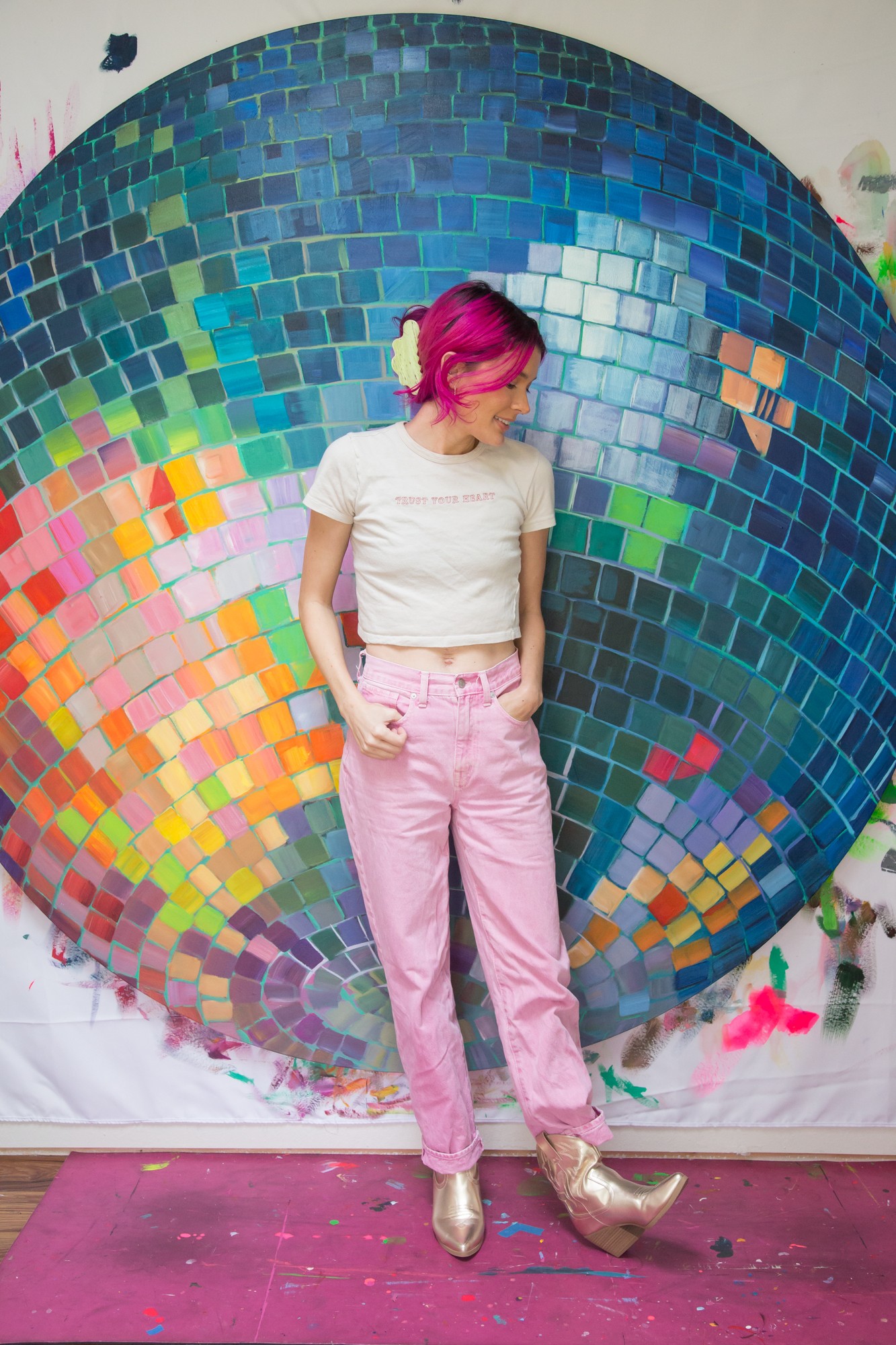 Person with pink hair in white shirt and pink pants stands in front of a colorful mosaic backdrop.
