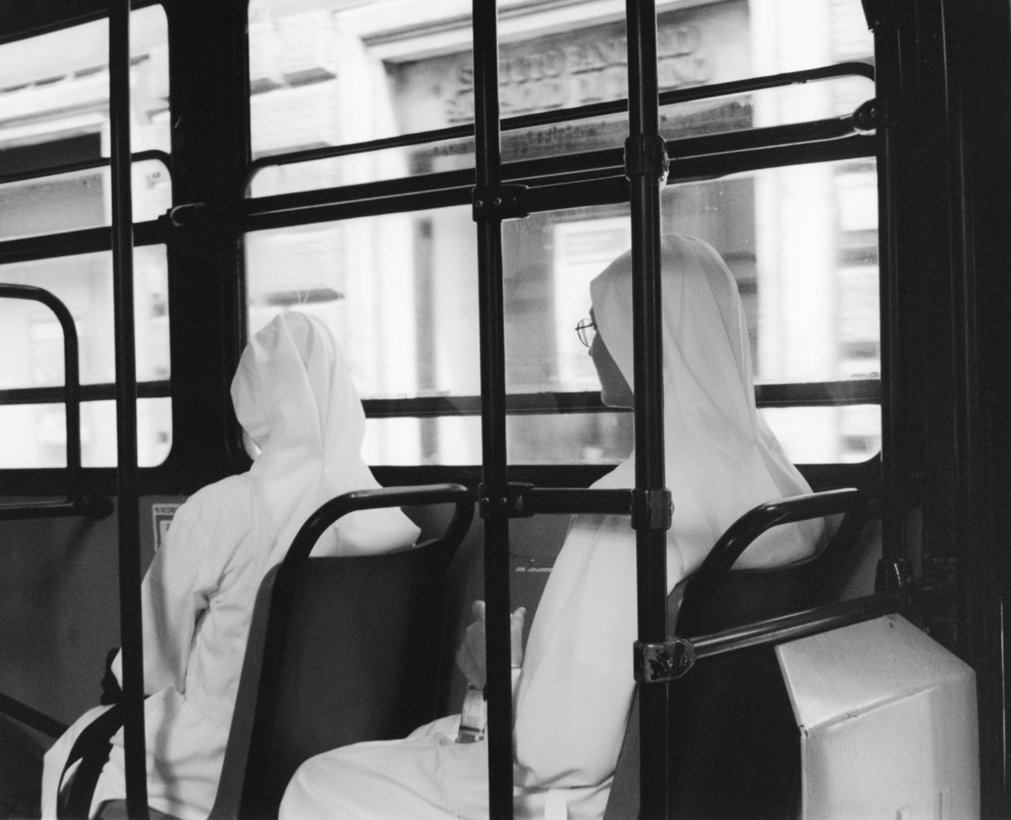 Two people wearing head coverings sit on a bus, viewed through window bars, with city buildings outside.