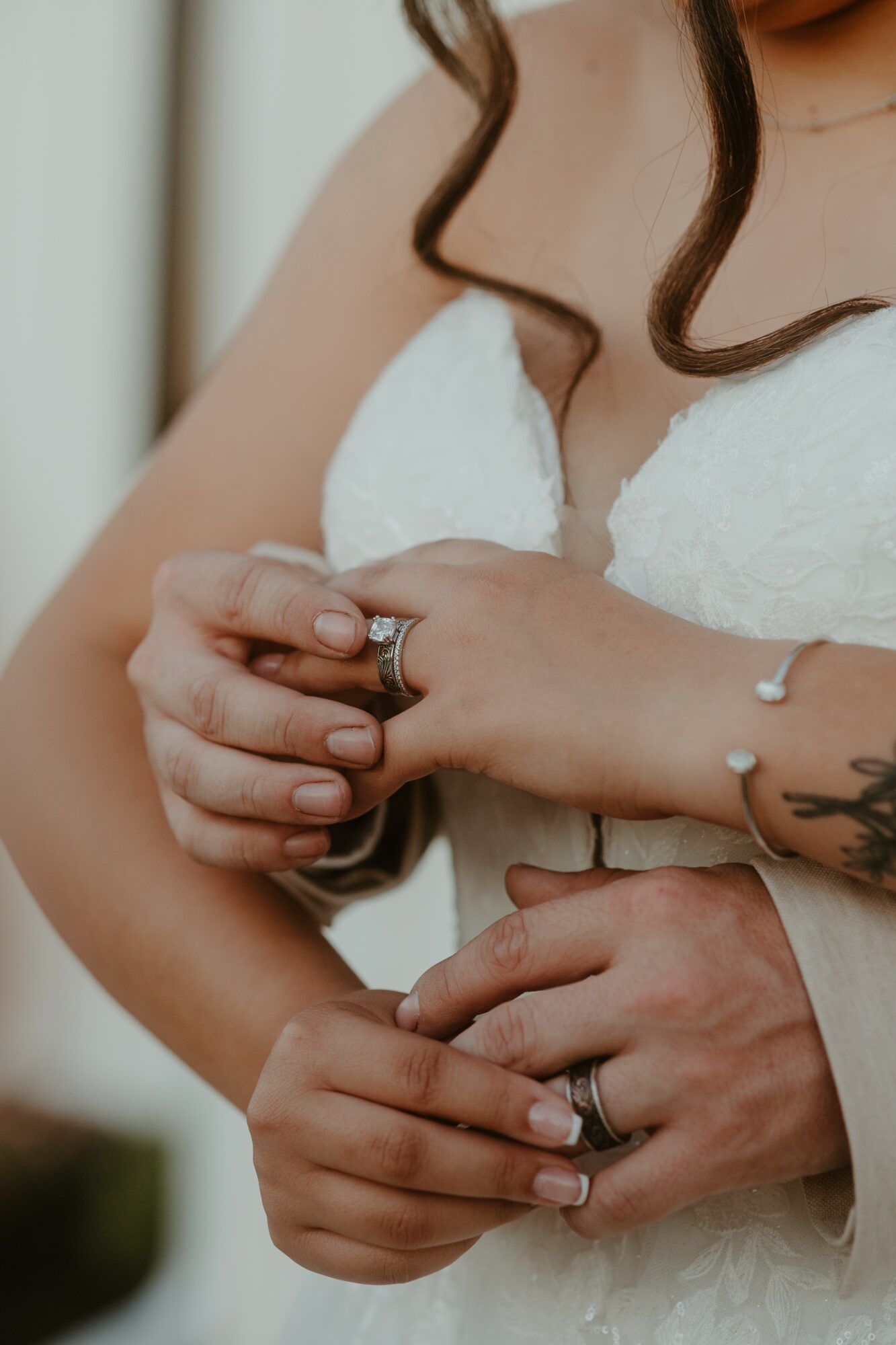 Close-up of a bride's hands holding a groom's hand, both wearing wedding rings, with part of her white wedding dress visible.