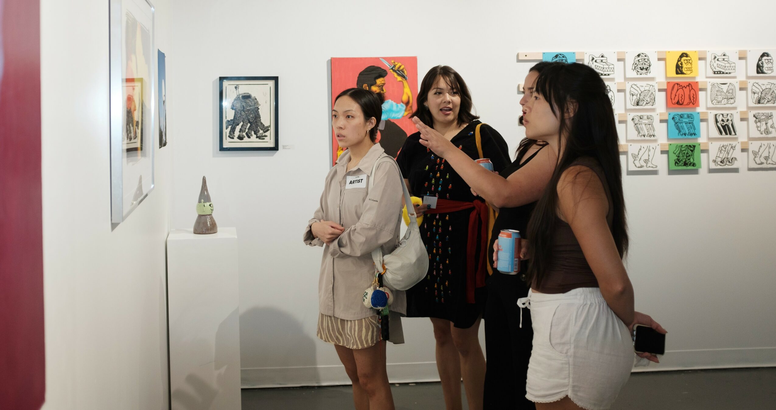 Three women in an art gallery looking at artwork, with colorful pieces on the wall behind them.