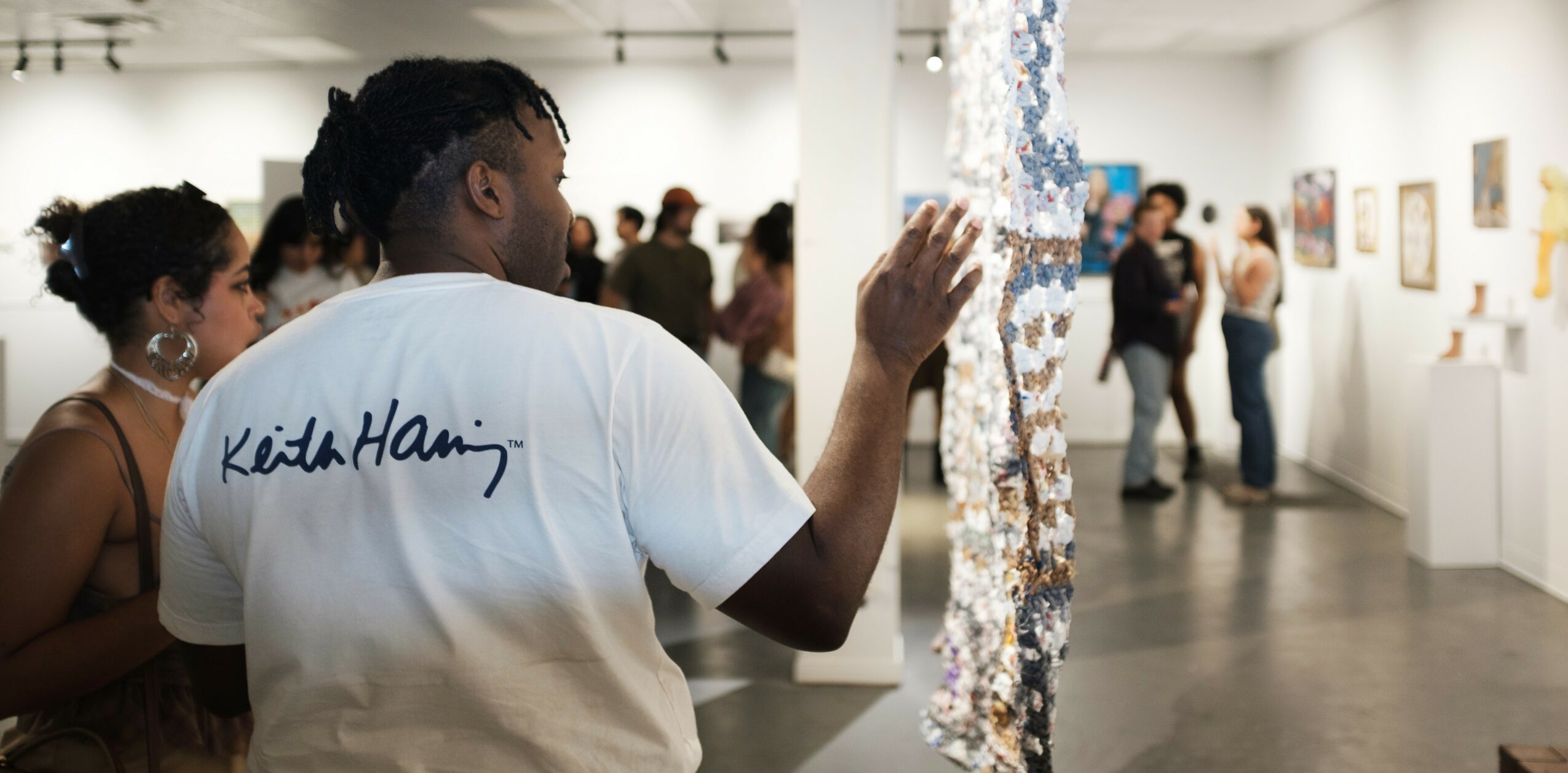 Person with braided hair and white T-shirt touching an art installation in gallery, surrounded by visitors.