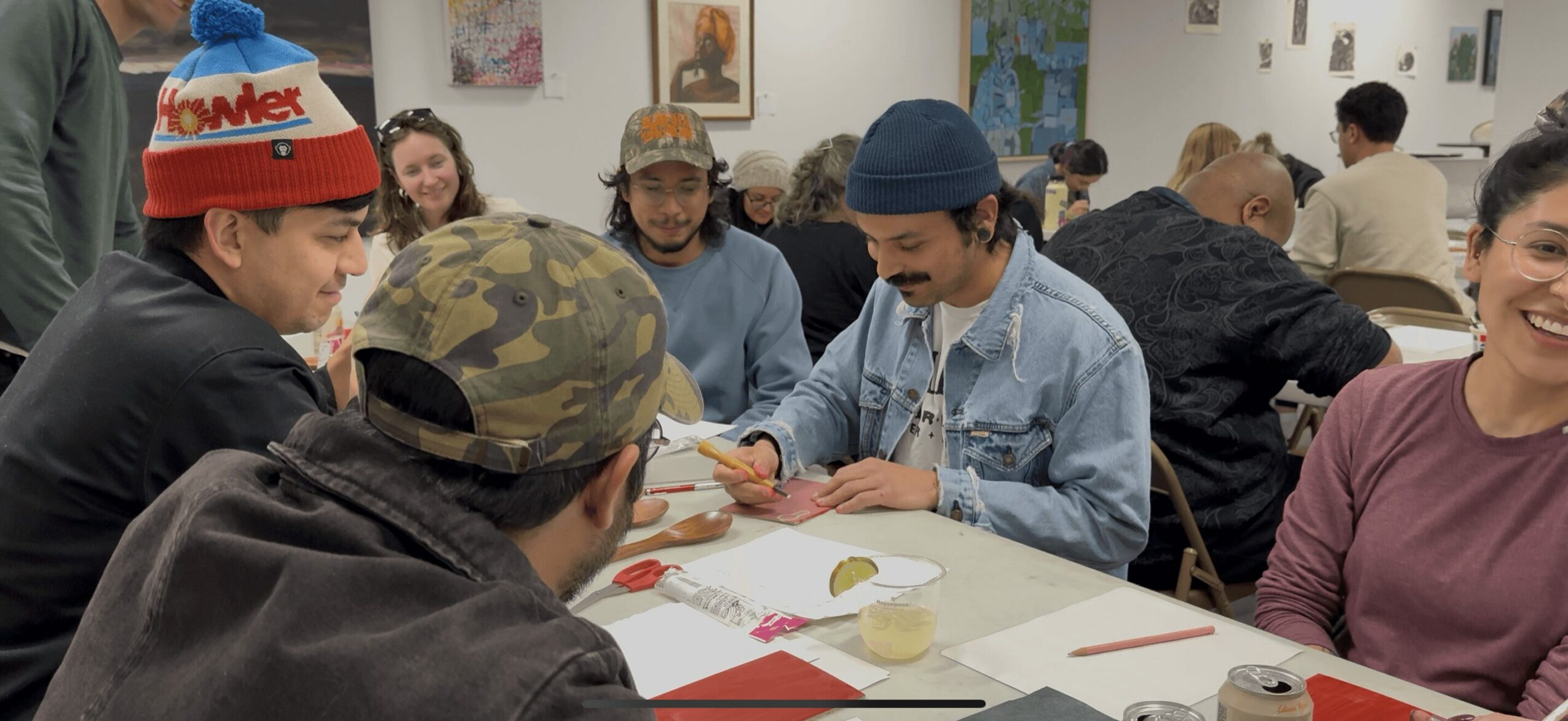 Group of young people sitting around a table, engaging in conversation and activities, in a room with artwork on the walls.