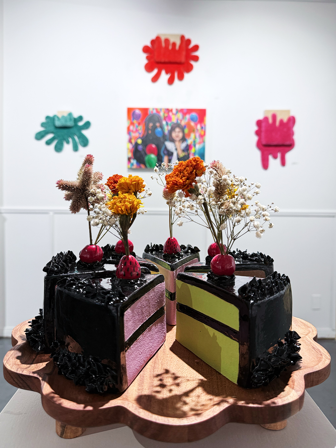 A black cake with pink and green layers, decorated with flowers and berries, on a wooden tray, with colorful wall art in background.