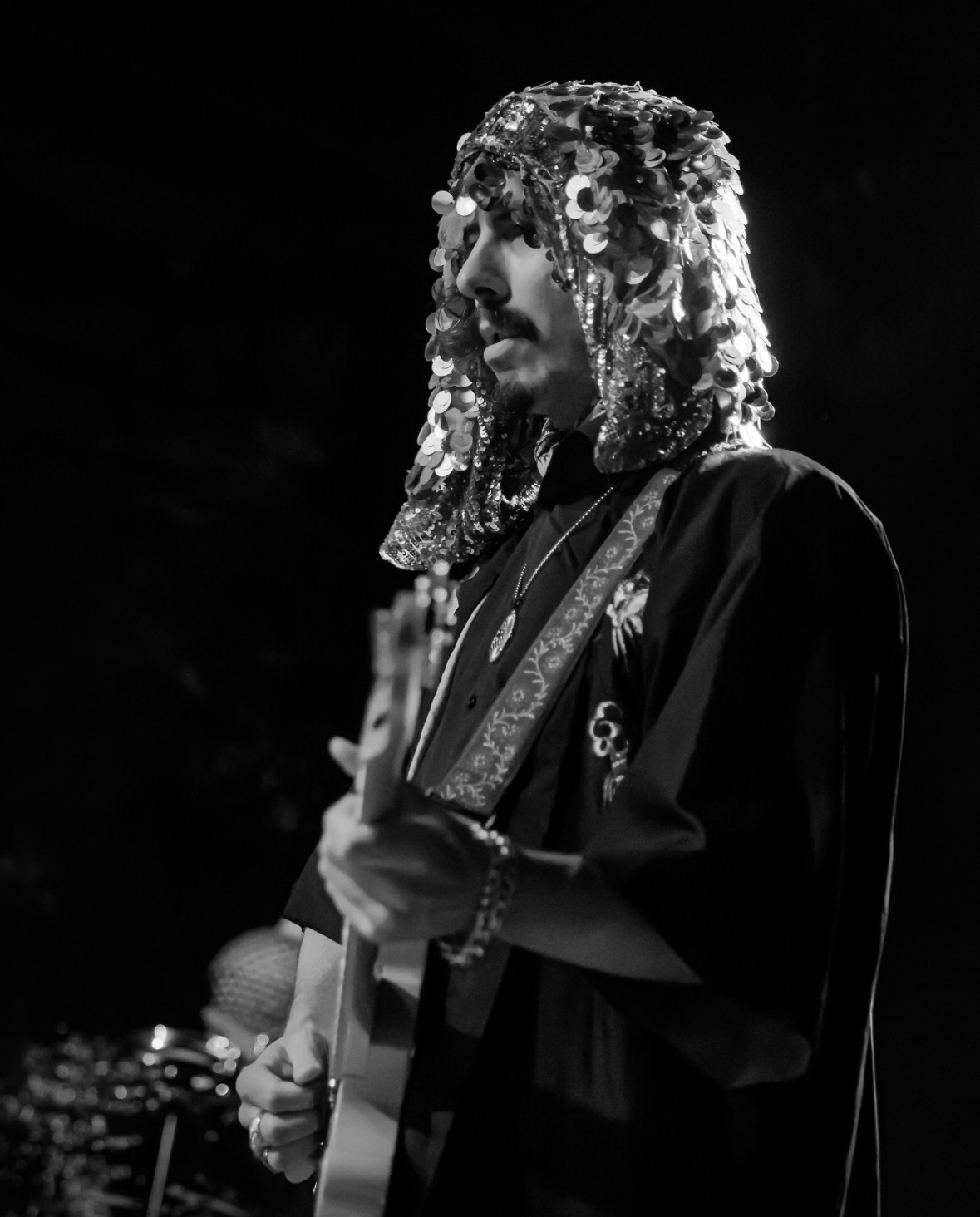 Person wearing a headdress with sequins, playing guitar on stage, in black and white.