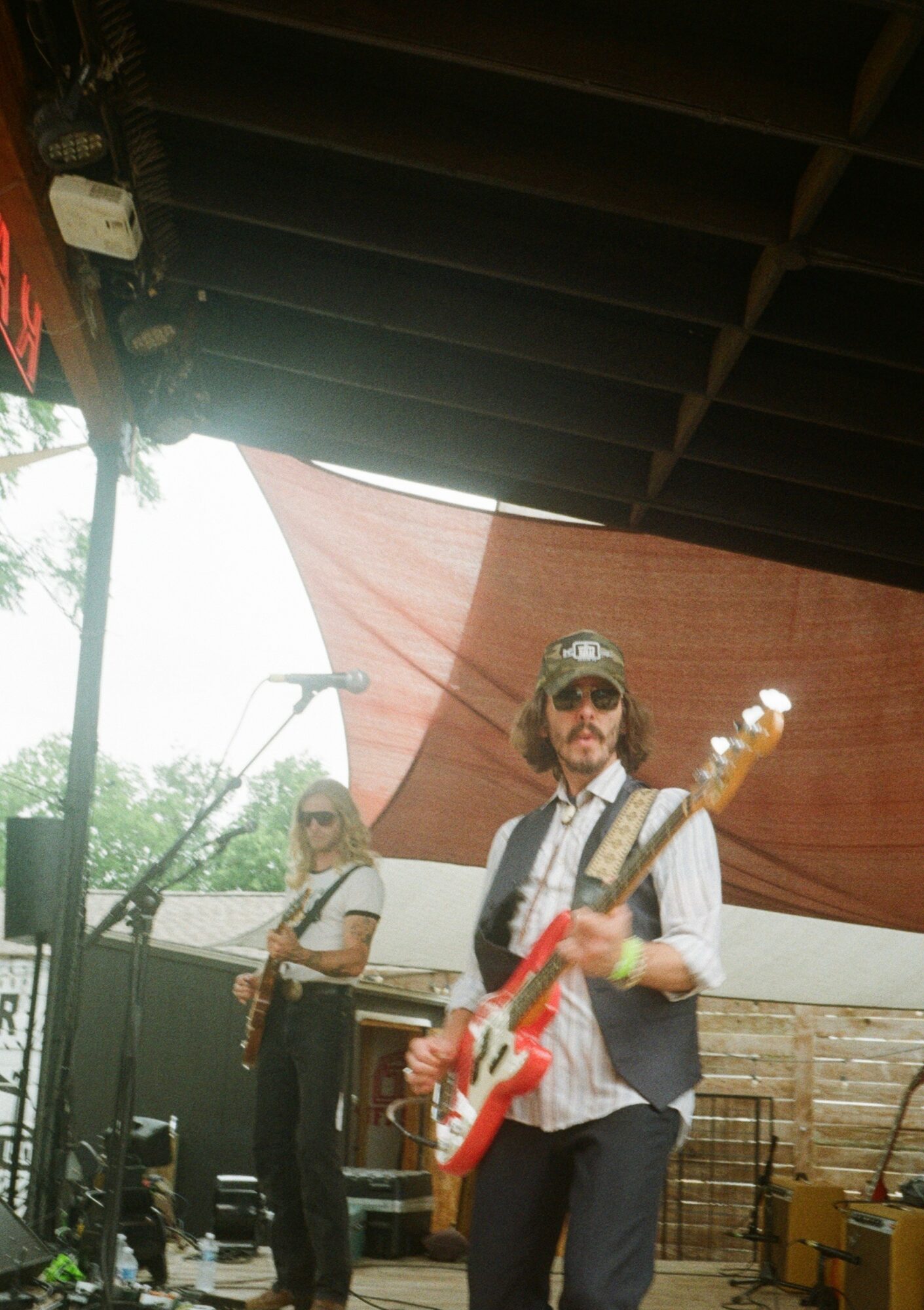 Two musicians performing outdoors under a canopy, one playing guitar and the other playing saxophone, with trees and sky in background.