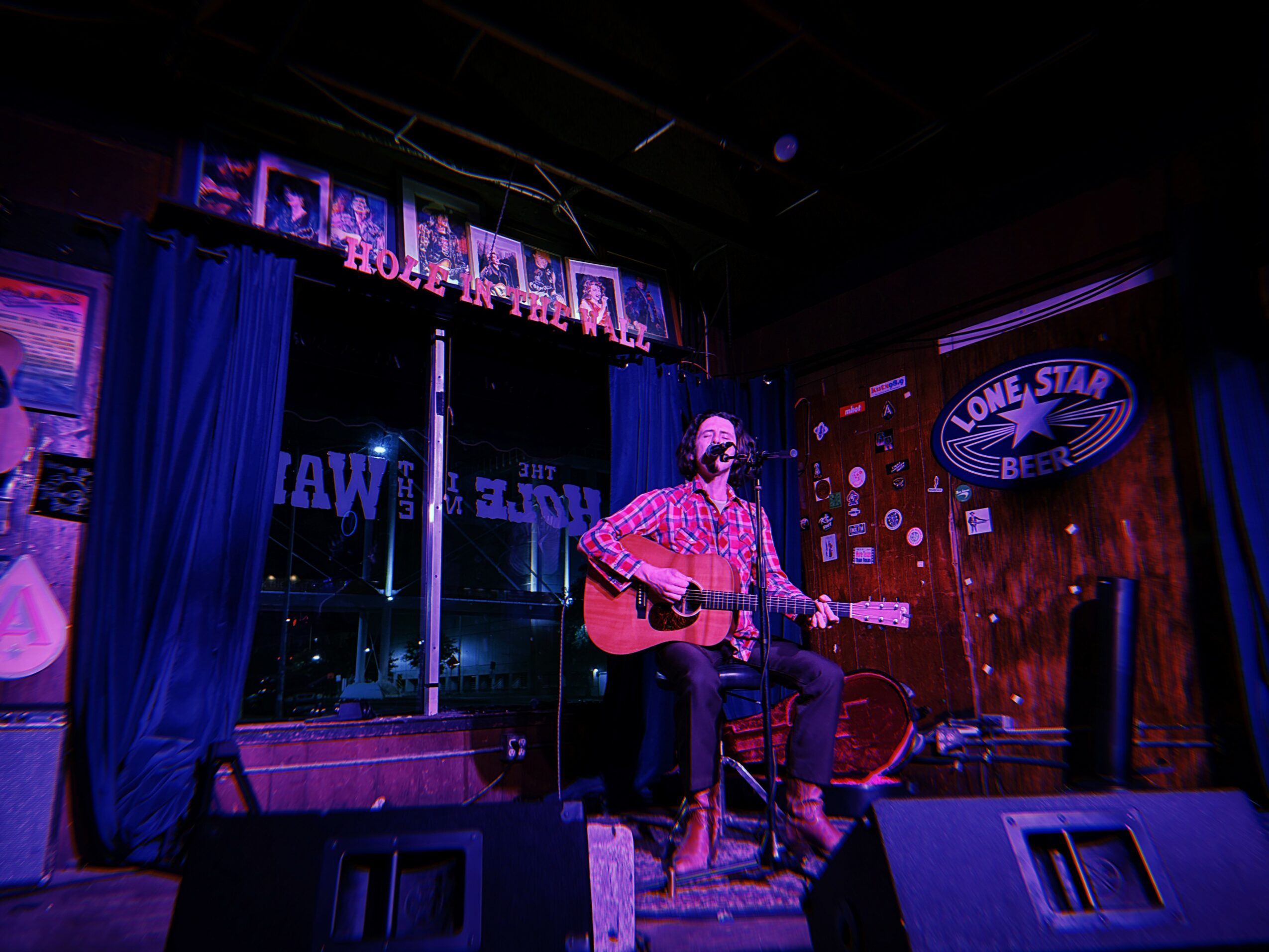 Person playing guitar on stage with neon signs and a red curtain background, in a dimly lit venue.