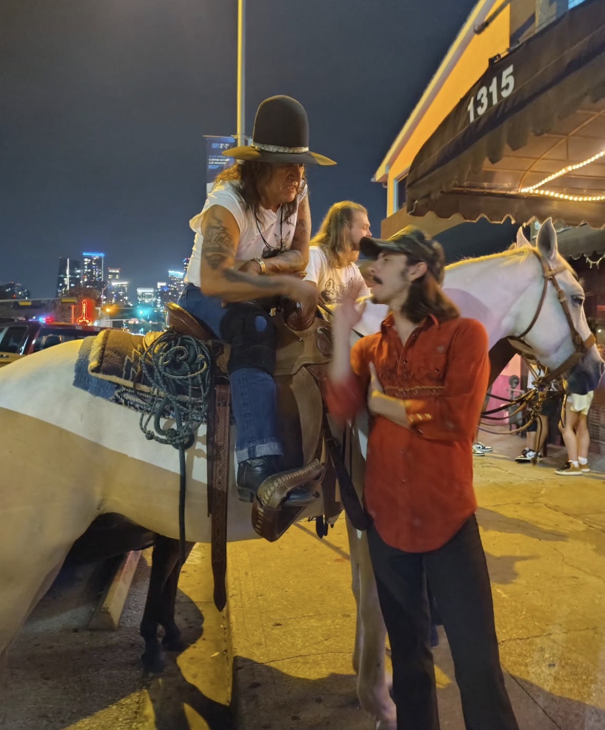 Two women and a horse at night, one woman sitting on the horse and the other standing nearby, city lights in background.