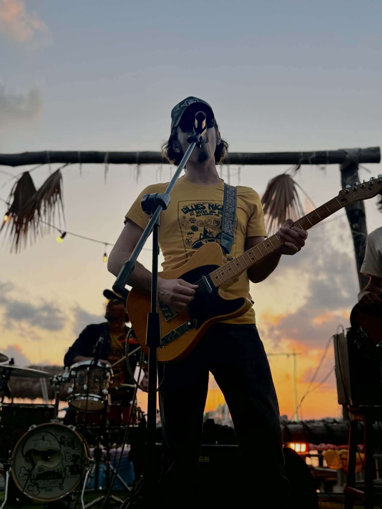 Person singing into microphone and playing guitar during sunset, with drummer in background and outdoor setting.