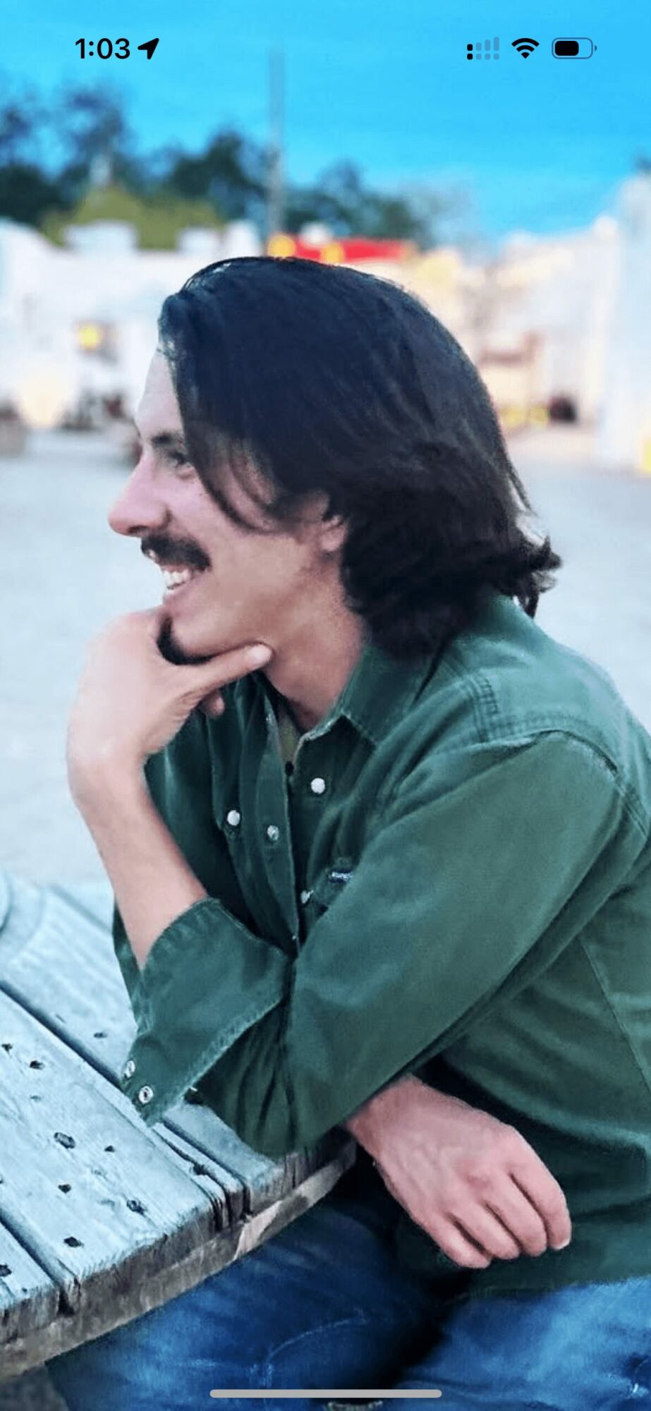 Person with dark hair and mustache sitting outdoors, smiling, wearing a green shirt, resting chin on hand, leaning on a wooden surface.