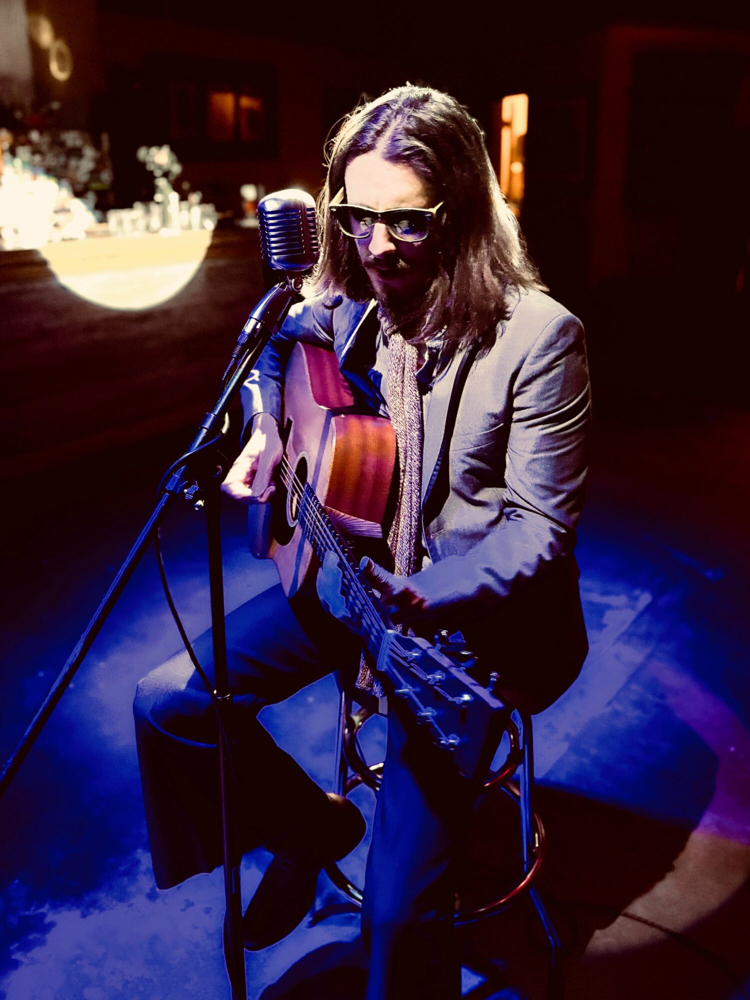 Person with long hair, sunglasses, gray blazer, playing guitar and singing into a microphone on stage.