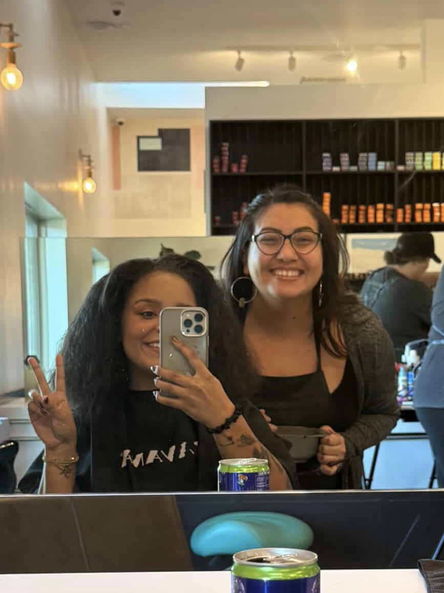 Two women smiling and taking a selfie in a cafe, one making a peace sign, shelves with books behind them.