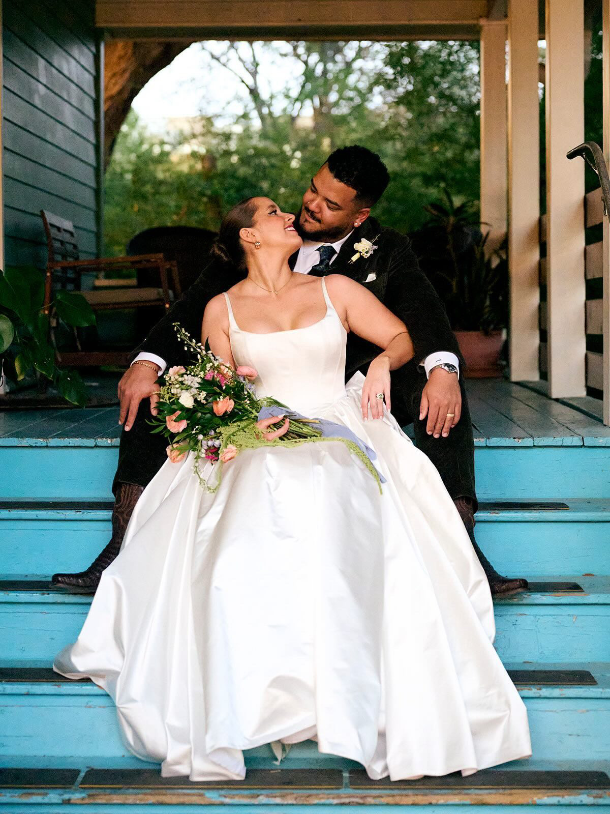 Bride and groom sitting on steps, smiling and looking at each other, bride holding a bouquet, outdoor setting