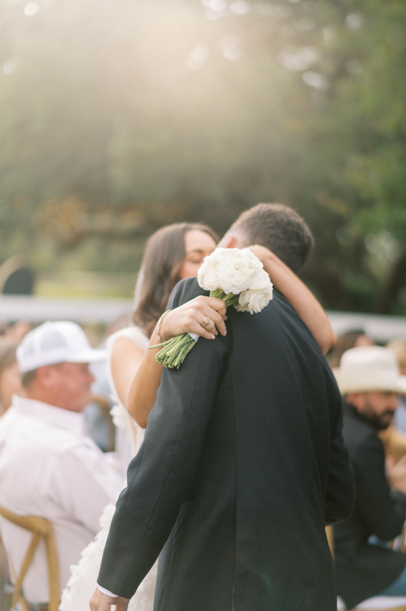 A couple embraces outdoors, with the woman holding a bouquet of white flowers, surrounded by people and trees.