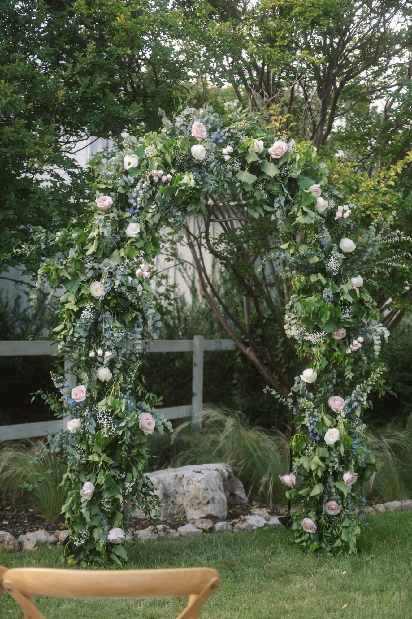 Flower arch with pink and white roses and greenery outdoors, trees and a fence in background.