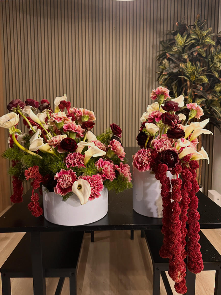 Two large floral arrangements in white round vases with pink, white, and dark purple flowers on a black table, background with vertical striped wall.