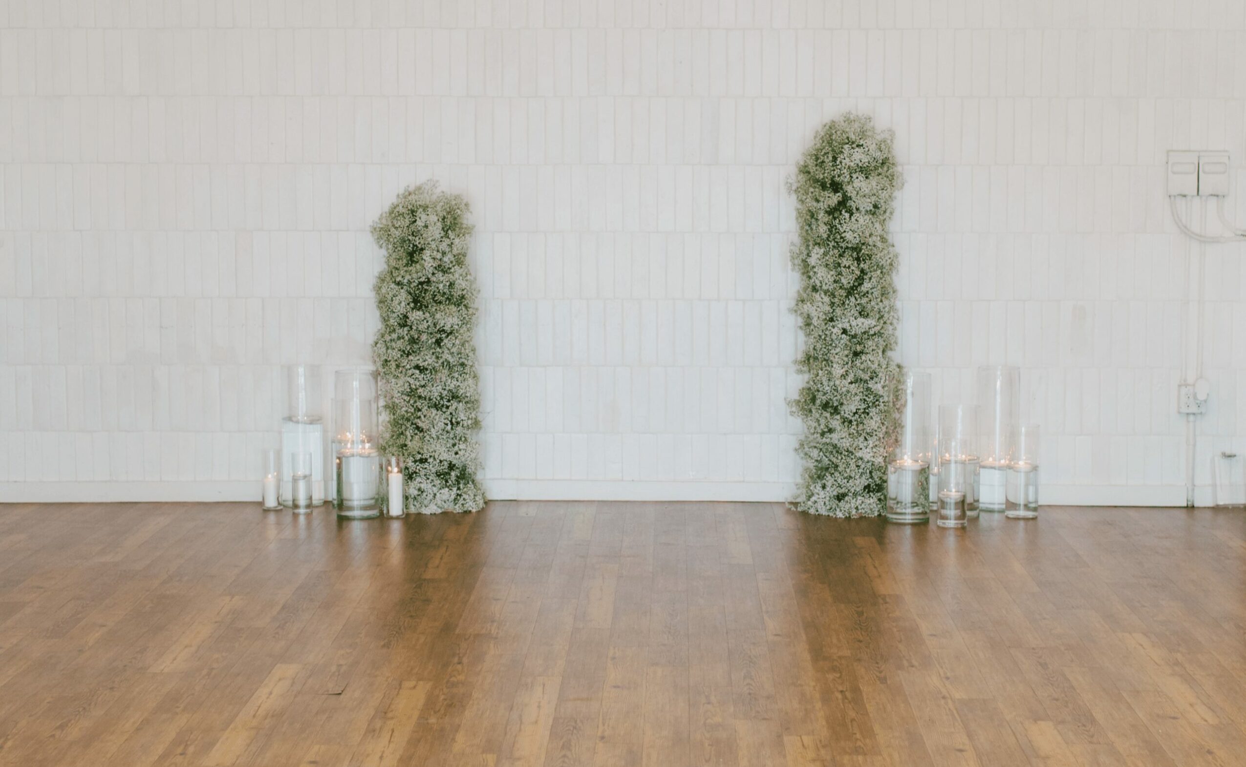 Two tall green plants with white flowers are placed against a white wall, with candles in glass holders on the floor.