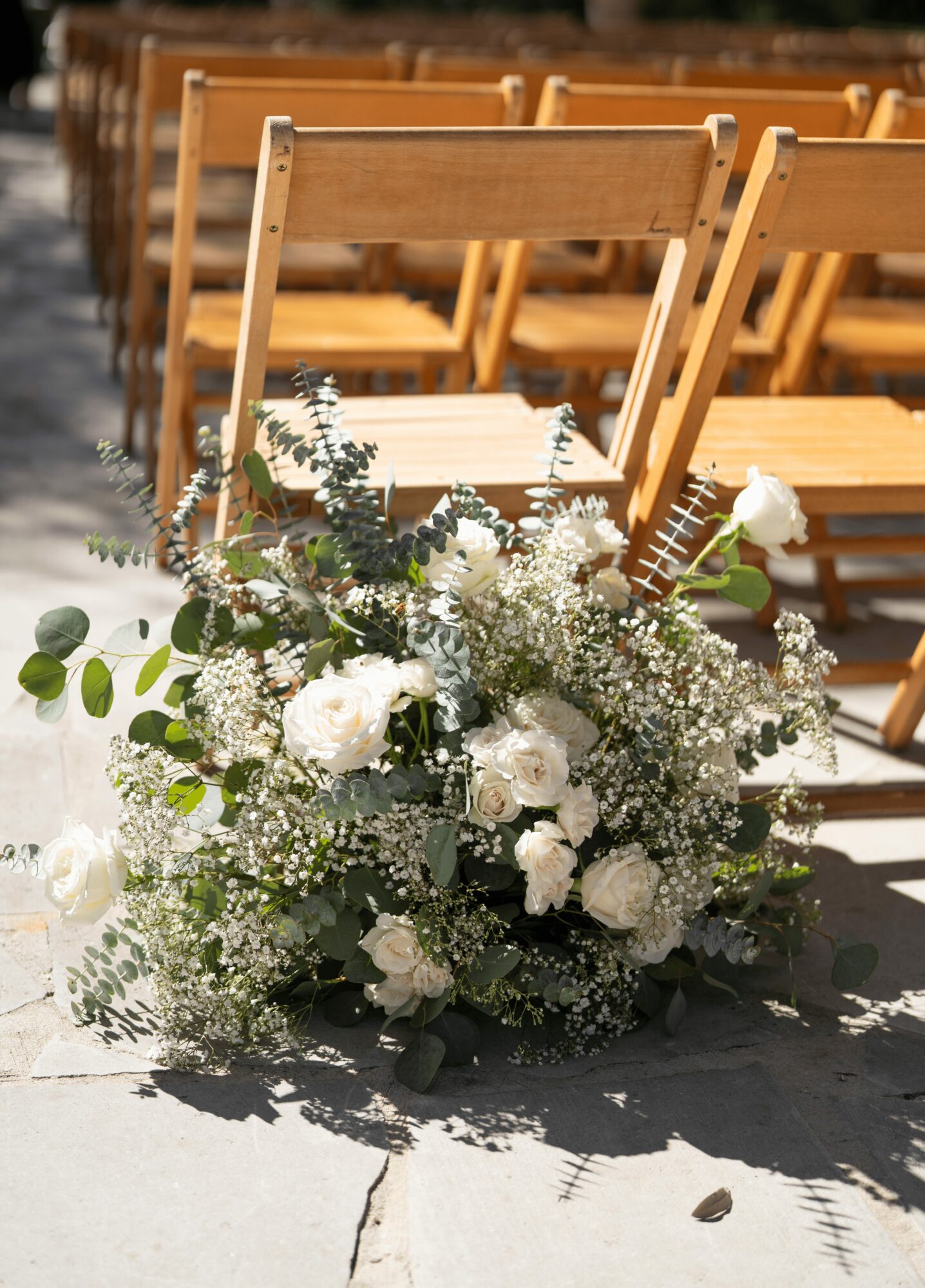 White floral arrangement with greenery on ground, chairs in background, outdoor setting, sunlight, shadows, concrete surface.