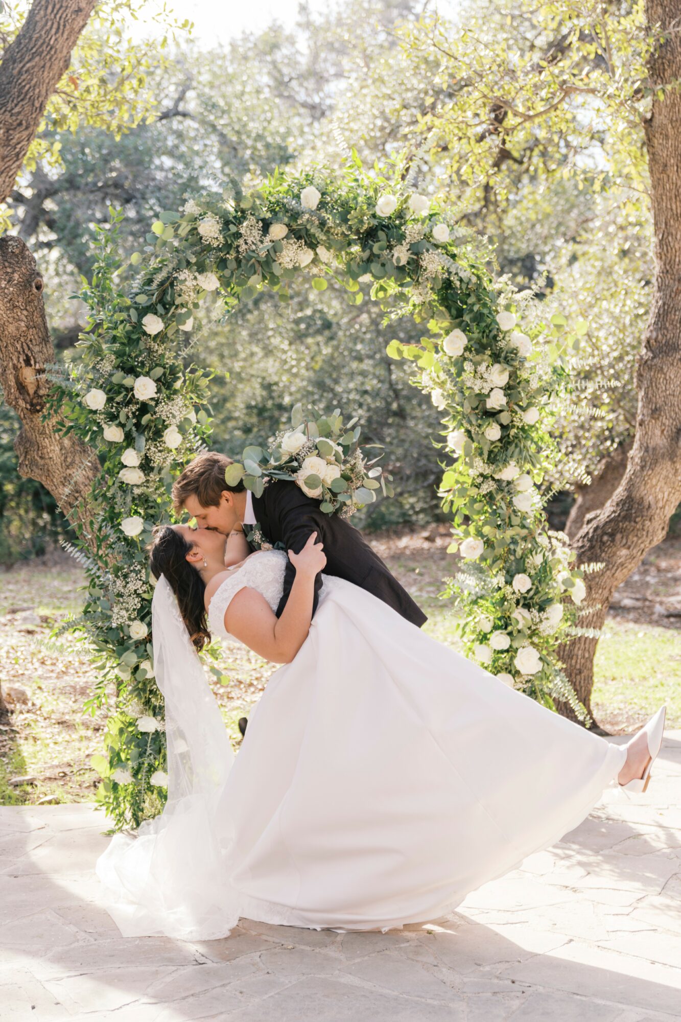 A bride and groom kiss outdoors under a floral arch, surrounded by trees and sunlight.