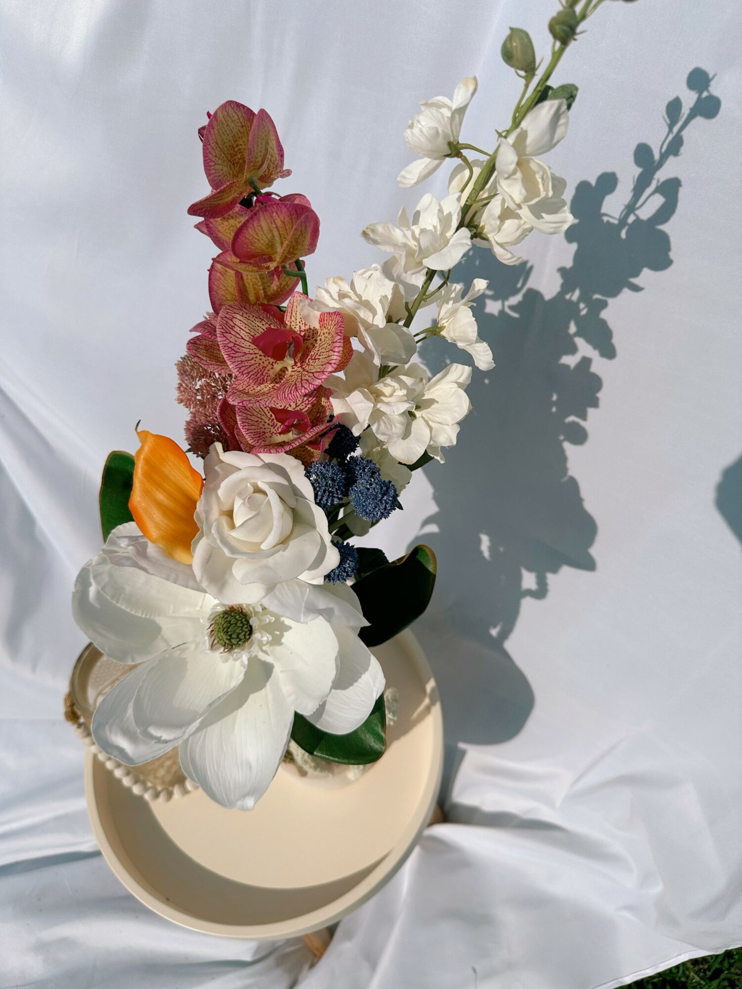 Flower arrangement with white, pink, orange, and dark blue flowers in a round vase, casting a shadow on a white background.