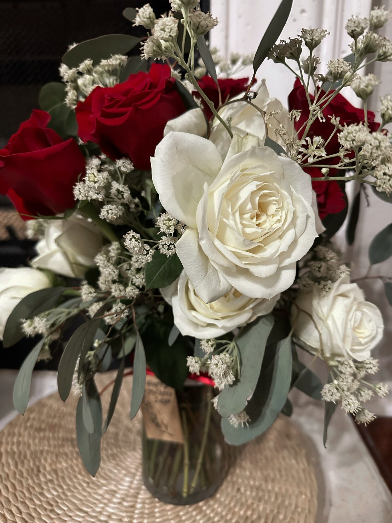 Bouquet of white roses, red roses, baby's breath, and greenery in a glass vase on a woven mat.