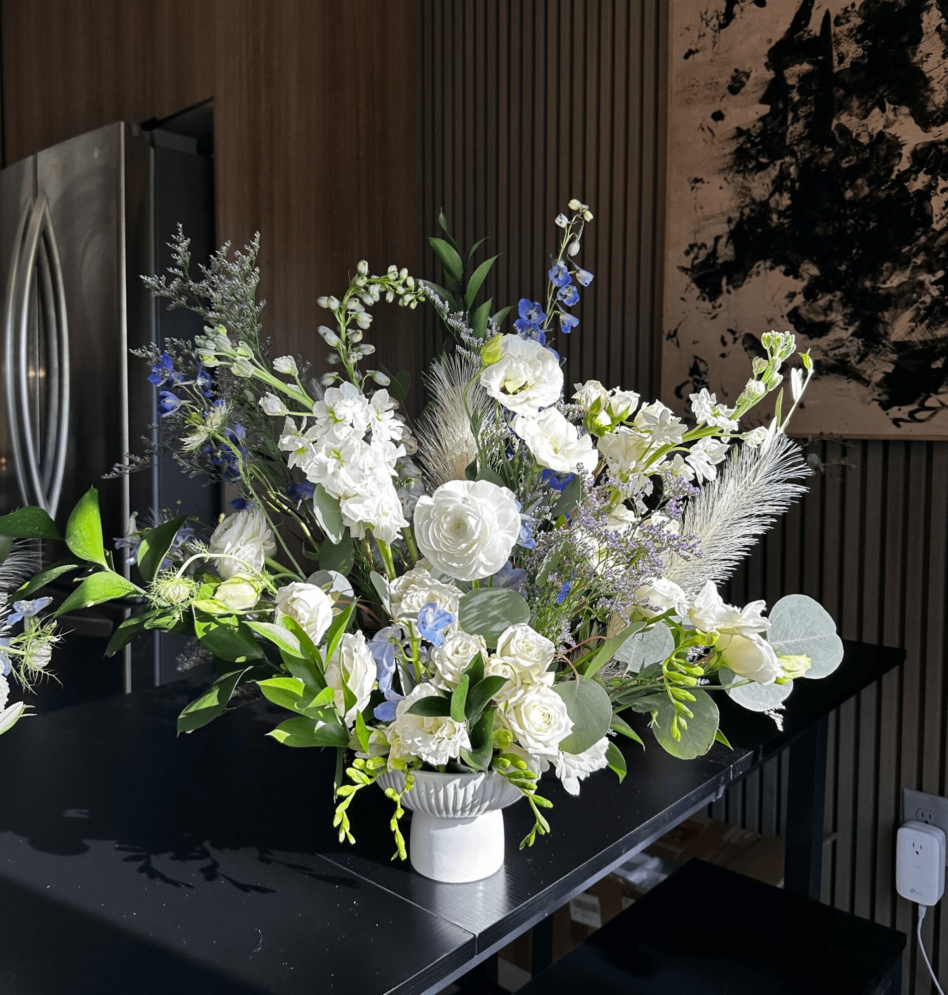 Large floral arrangement with white, blue, and green flowers in a white vase on a black table, indoors.