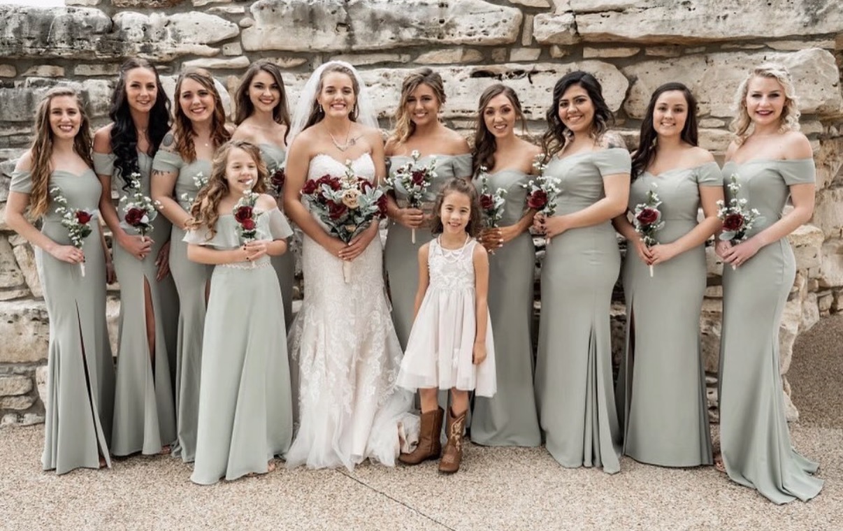 Group of women and children in formal dresses, standing outdoors against a stone wall, smiling, with some holding bouquets.