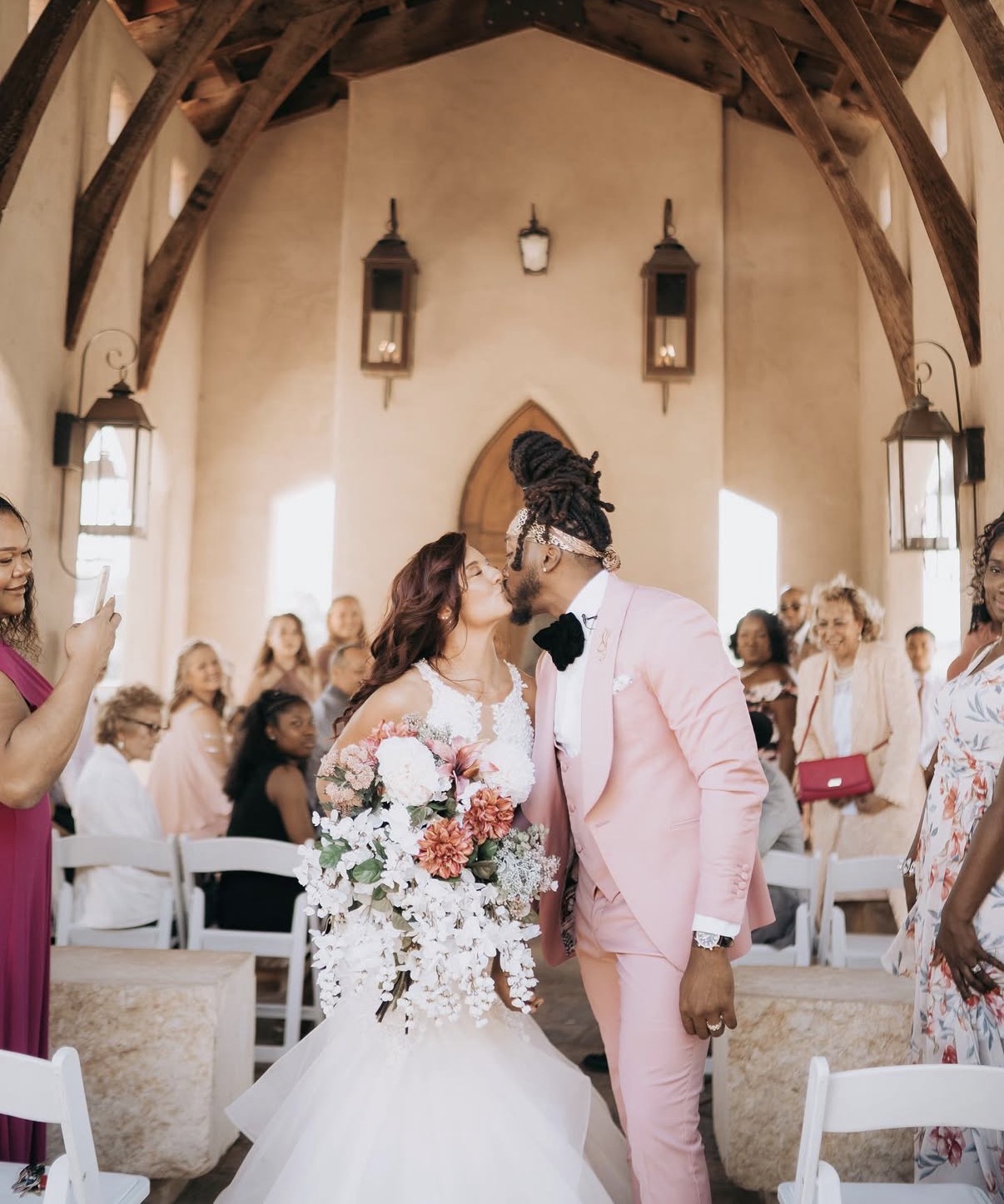 Bride and groom kiss at altar during wedding ceremony, guests seated and standing around them, church interior with wooden beams and lanterns.