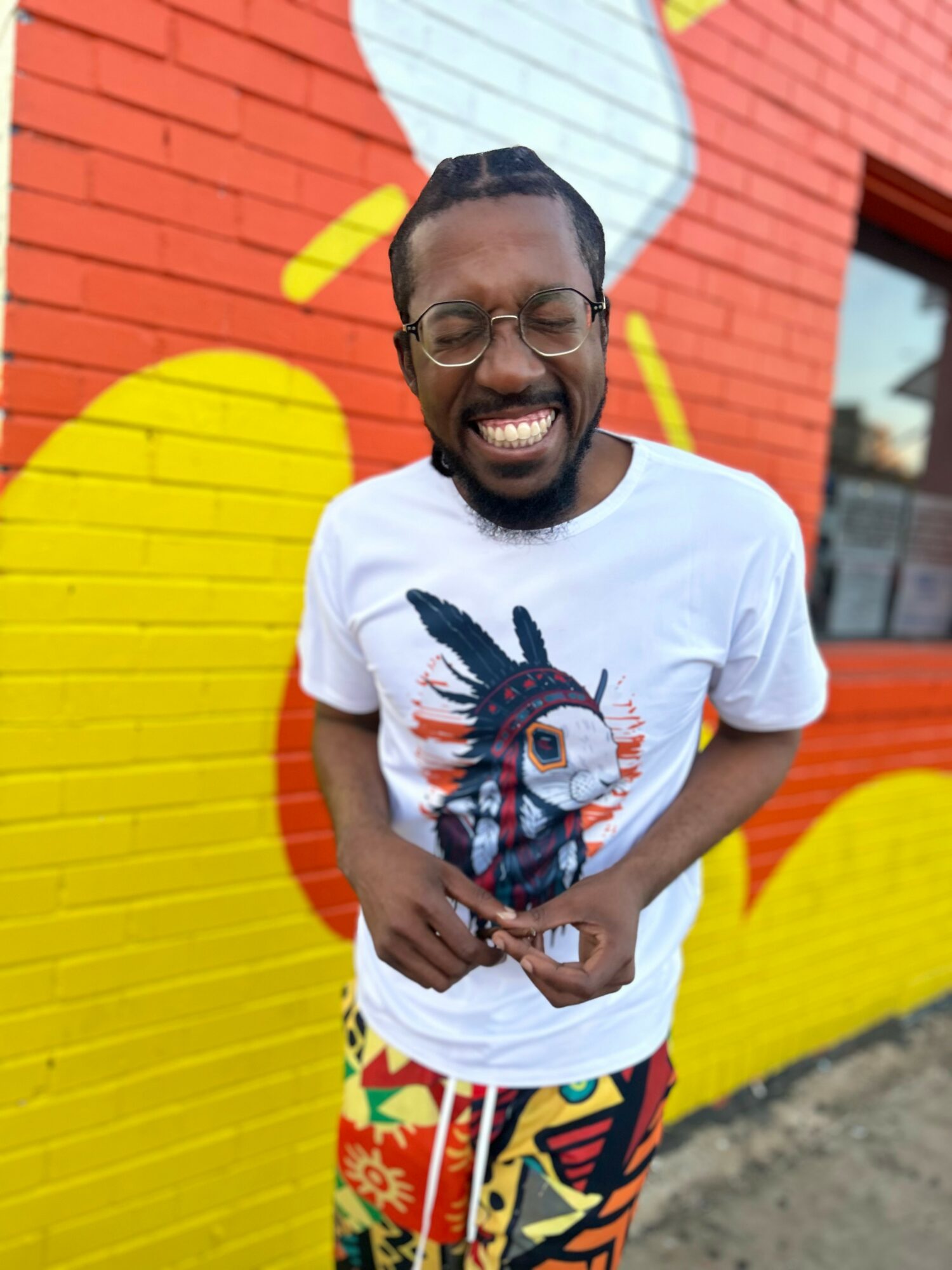 Smiling man with glasses and a beard stands in front of a colorful mural, wearing a white t-shirt with a graphic design.