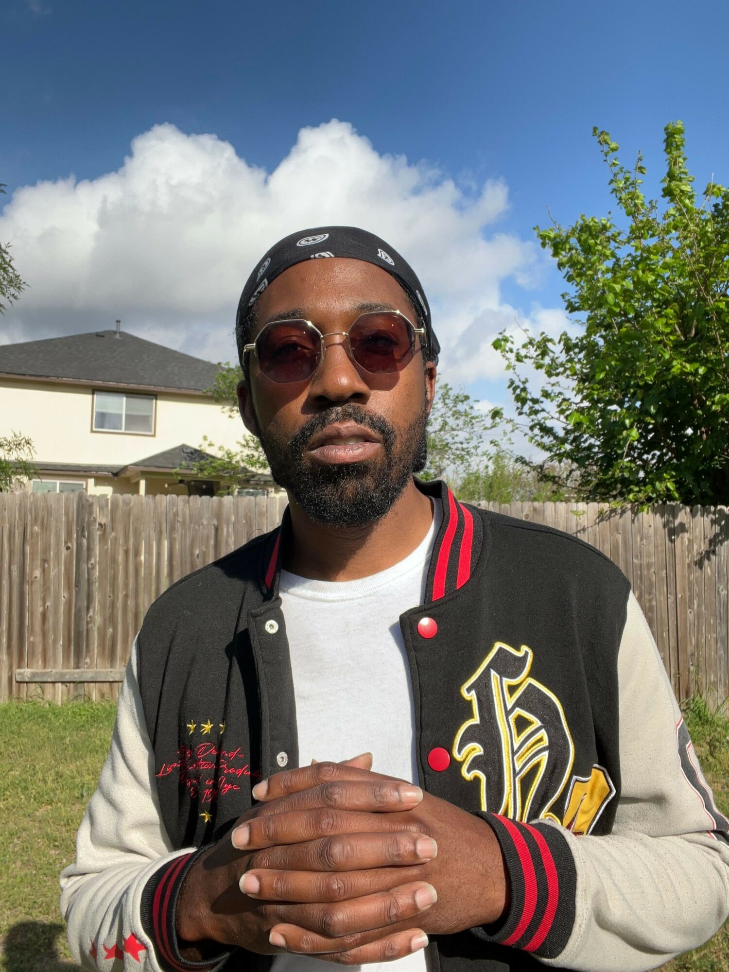 Man wearing sunglasses and a varsity jacket, standing outdoors with a wooden fence and house in background.