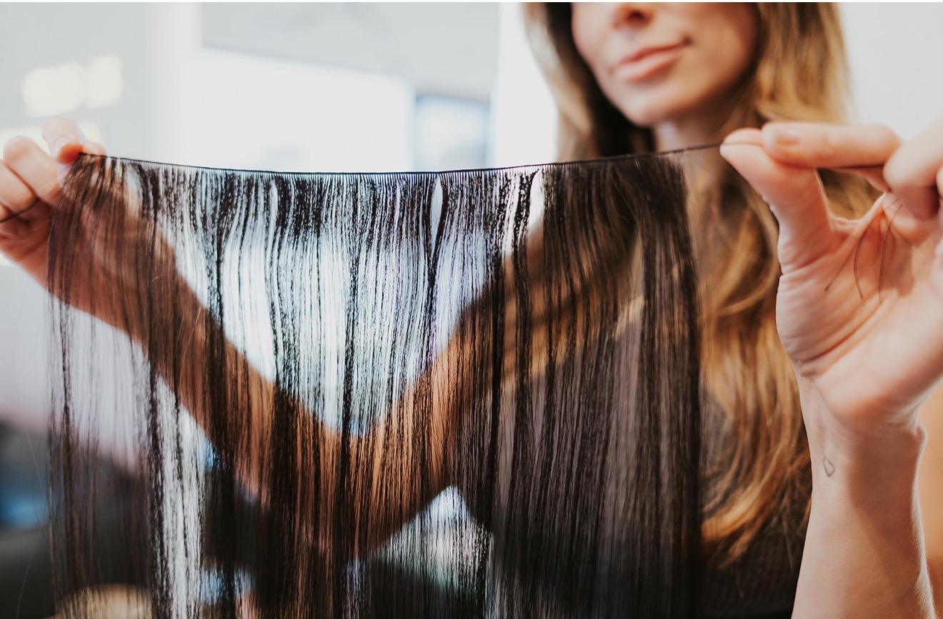 Person holding a transparent, black hair extension or wig, with a blurred background and a woman in the background.
