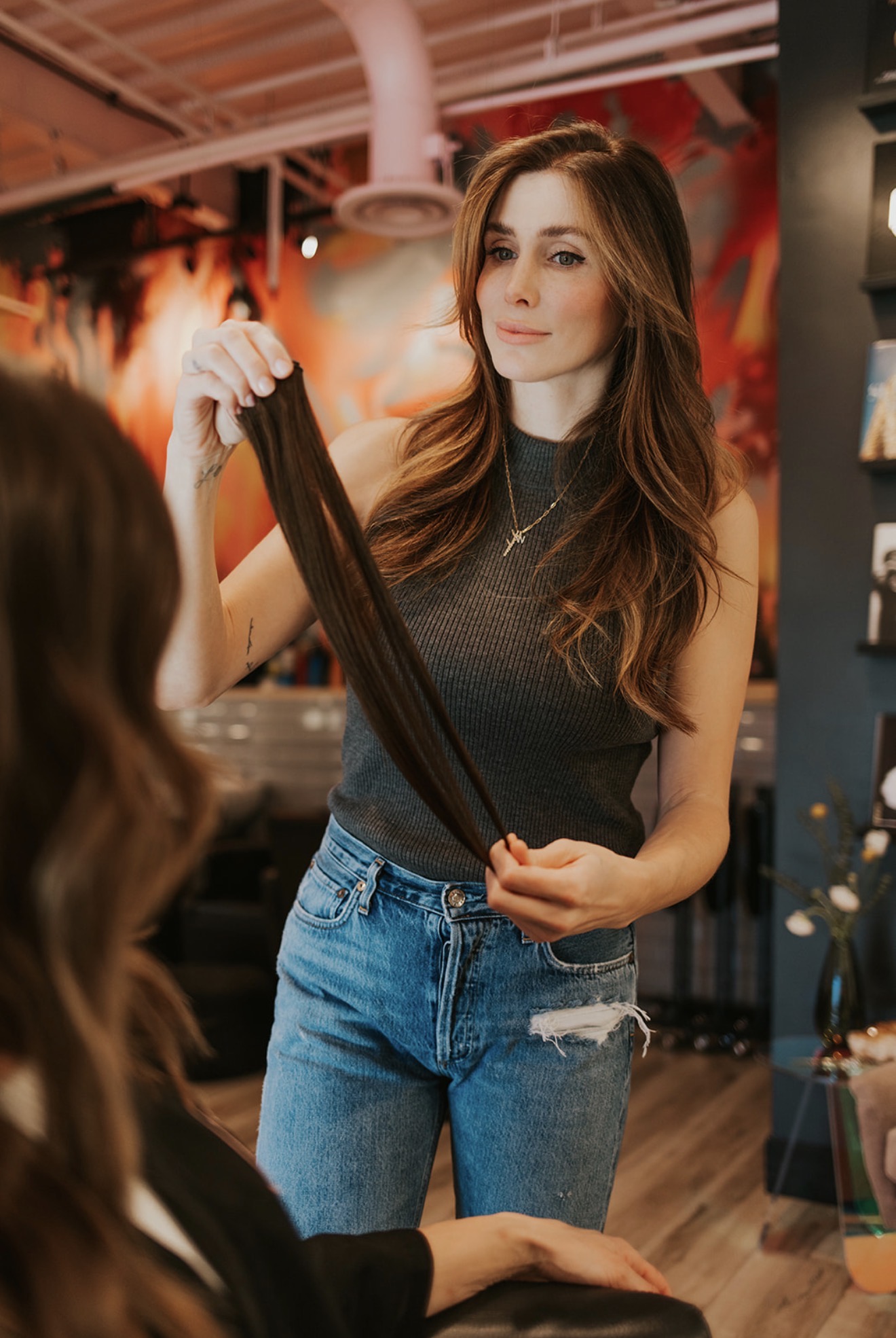 Woman with long brown hair holding a hair extension, wearing a sleeveless gray top and ripped jeans, in a salon or studio.
