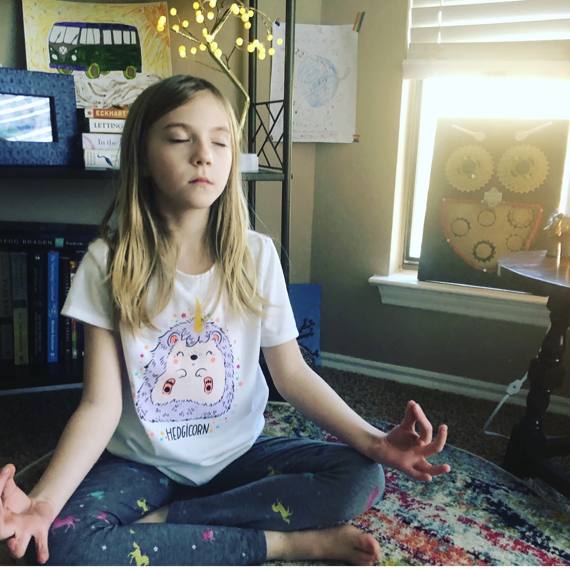 Girl sitting cross-legged on a colorful rug, meditating with eyes closed in a room with books and sunlight.