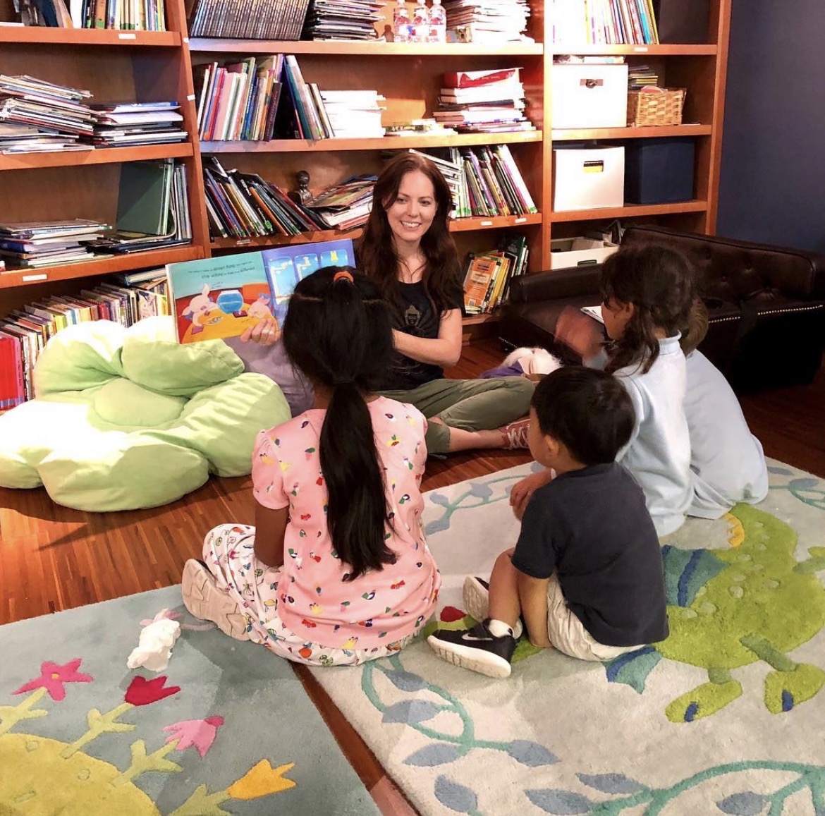Woman reading to children in a cozy room with bookshelves, colorful cushions, and a patterned rug.