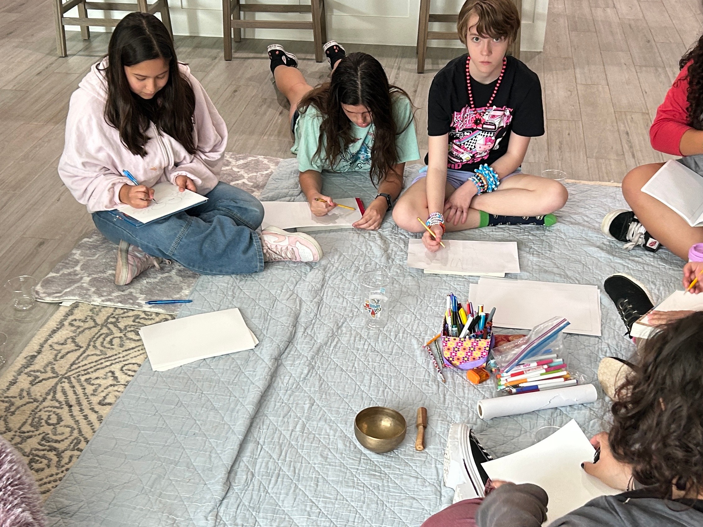 Four children sit on the floor around a table with art supplies, drawing and coloring. The table has a cloth and a small bowl.