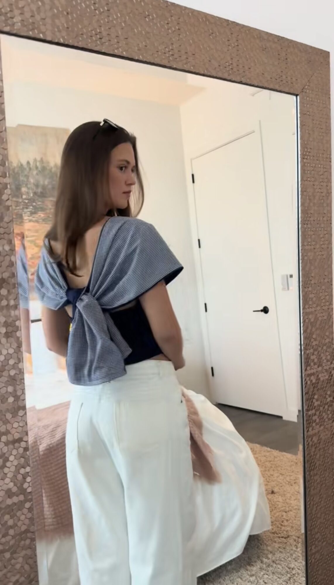 Young woman with shoulder-length hair stands in front of a mirror, wearing a blue top and white pants, in a bedroom.