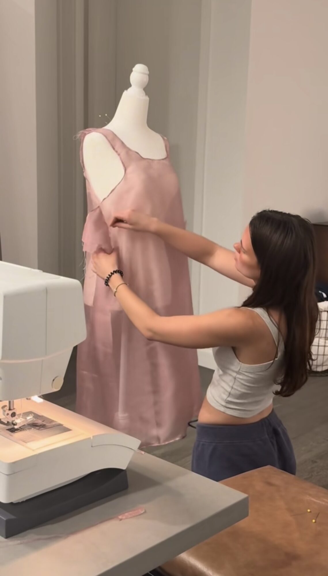 Person adjusting a pink dress on a mannequin in a room, with a desk and computer in the foreground.