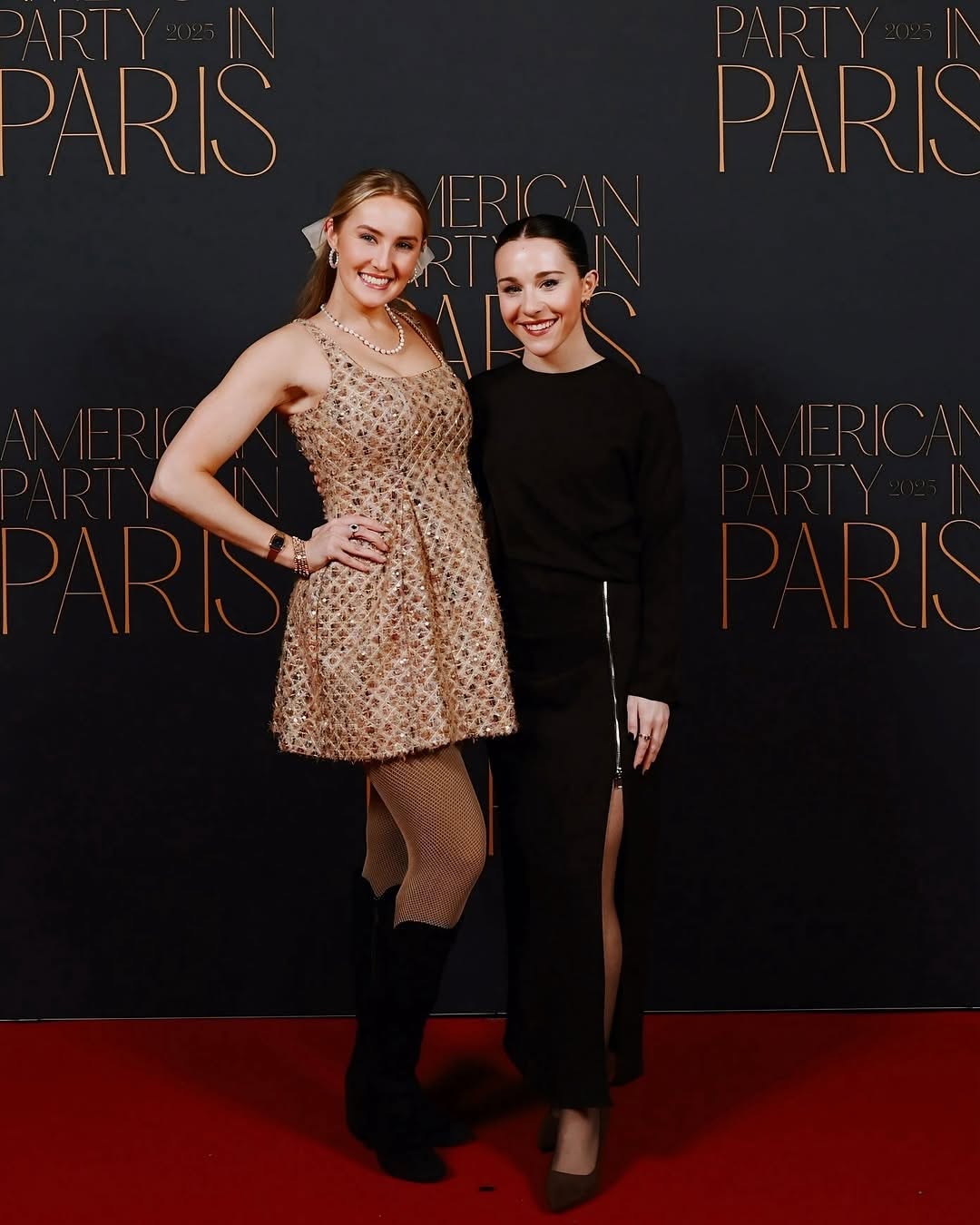 Two women stand together on a red carpet, smiling, in front of a black backdrop with gold text, one in a dress, one in black outfit.